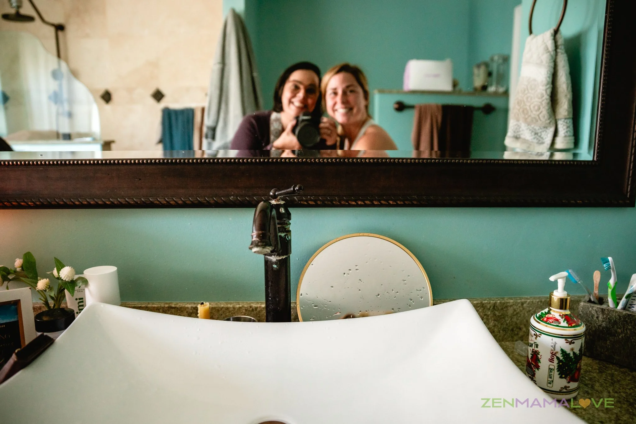 A large mirror above a bathroom sink reflects two smiling women, one with dark hair and the other with blonde hair, taking a selfie. Bathroom items like toothbrushes, a soap dispenser, and a decorative mug are on the counter, with towels hanging on the wall.