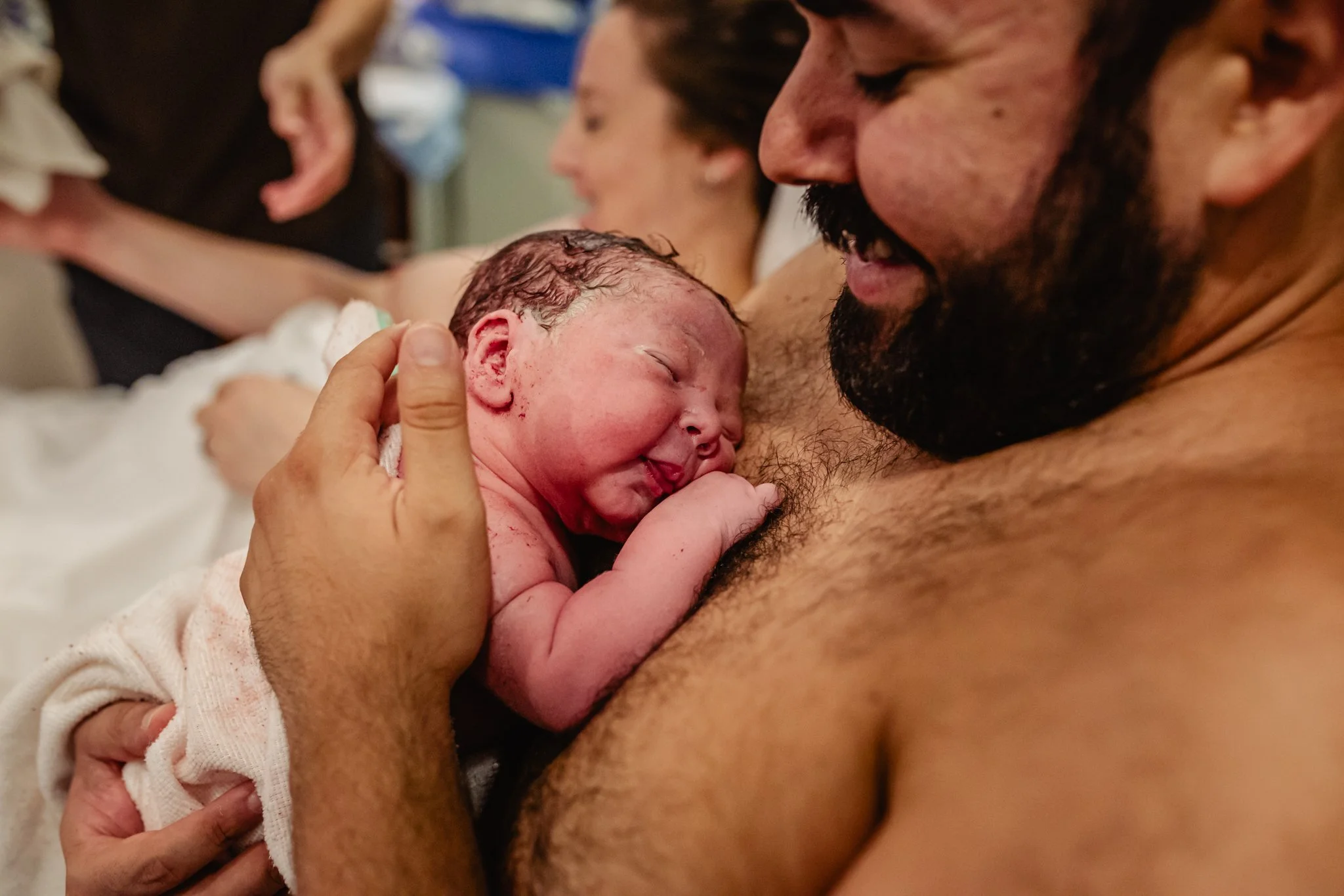 A bearded man holding a newborn baby close to his chest, with the baby's face resting on his chest. The scene appears to be in a hospital or birthing center, with other people in the background.