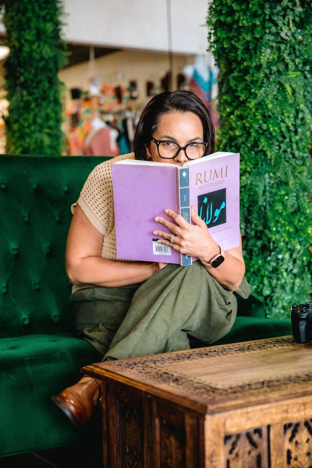 A woman with black hair, glasses, and a watch on her left wrist, sitting on a green couch, reading a purple book titled 'Rumi Illustrated' and winking at the camera.