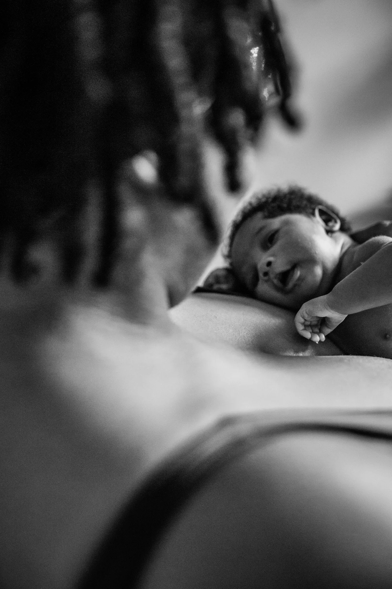 A black and white photo of a smiling baby lying on a surface, with their head resting on their arm, and an adult with dreadlocked hair looking at the baby in the background.