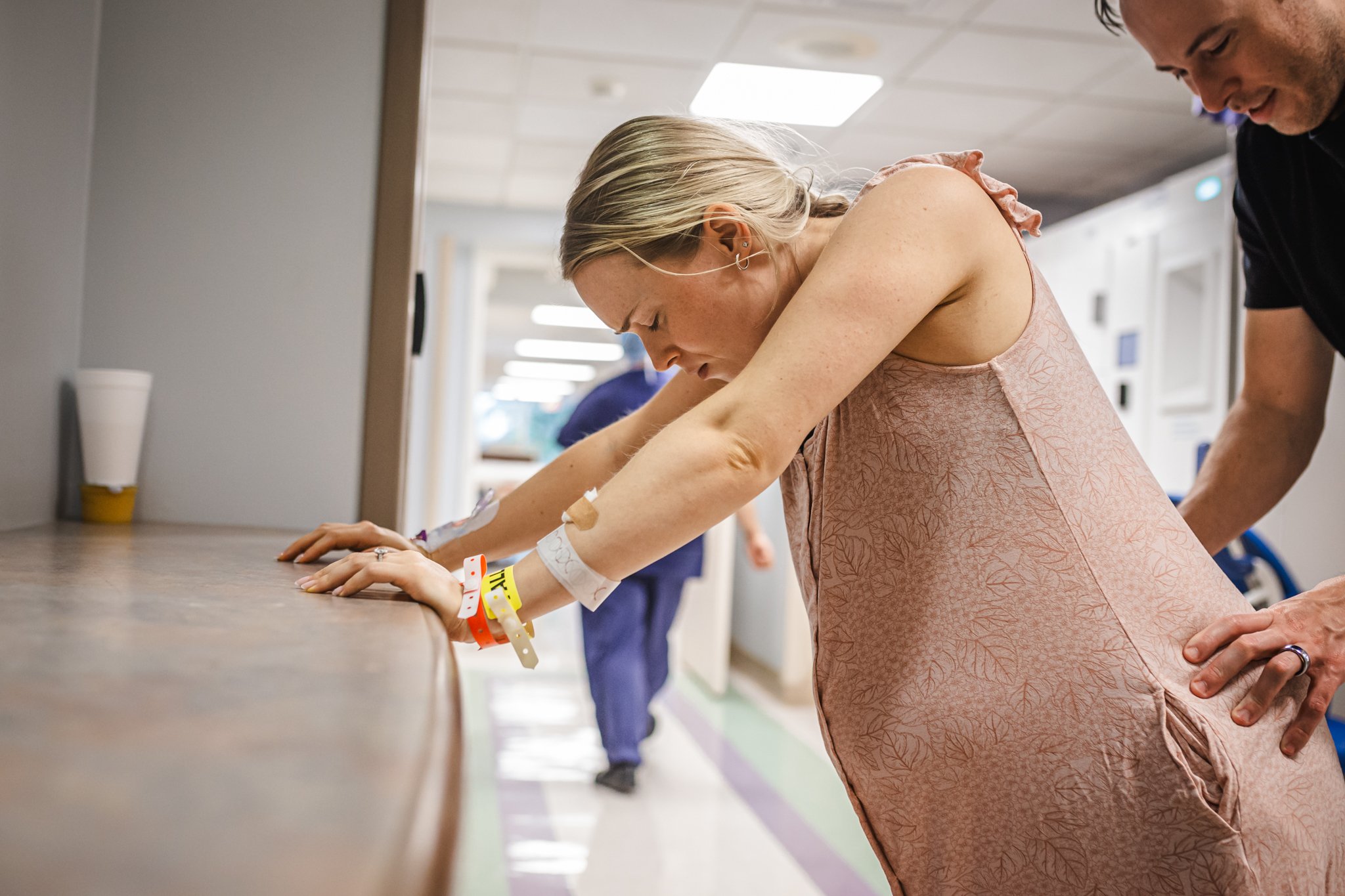 A woman leaning over a hospital bed with intravenous lines and medical wristbands, accompanied by a man in a black shirt standing nearby; they are in a hospital corridor.