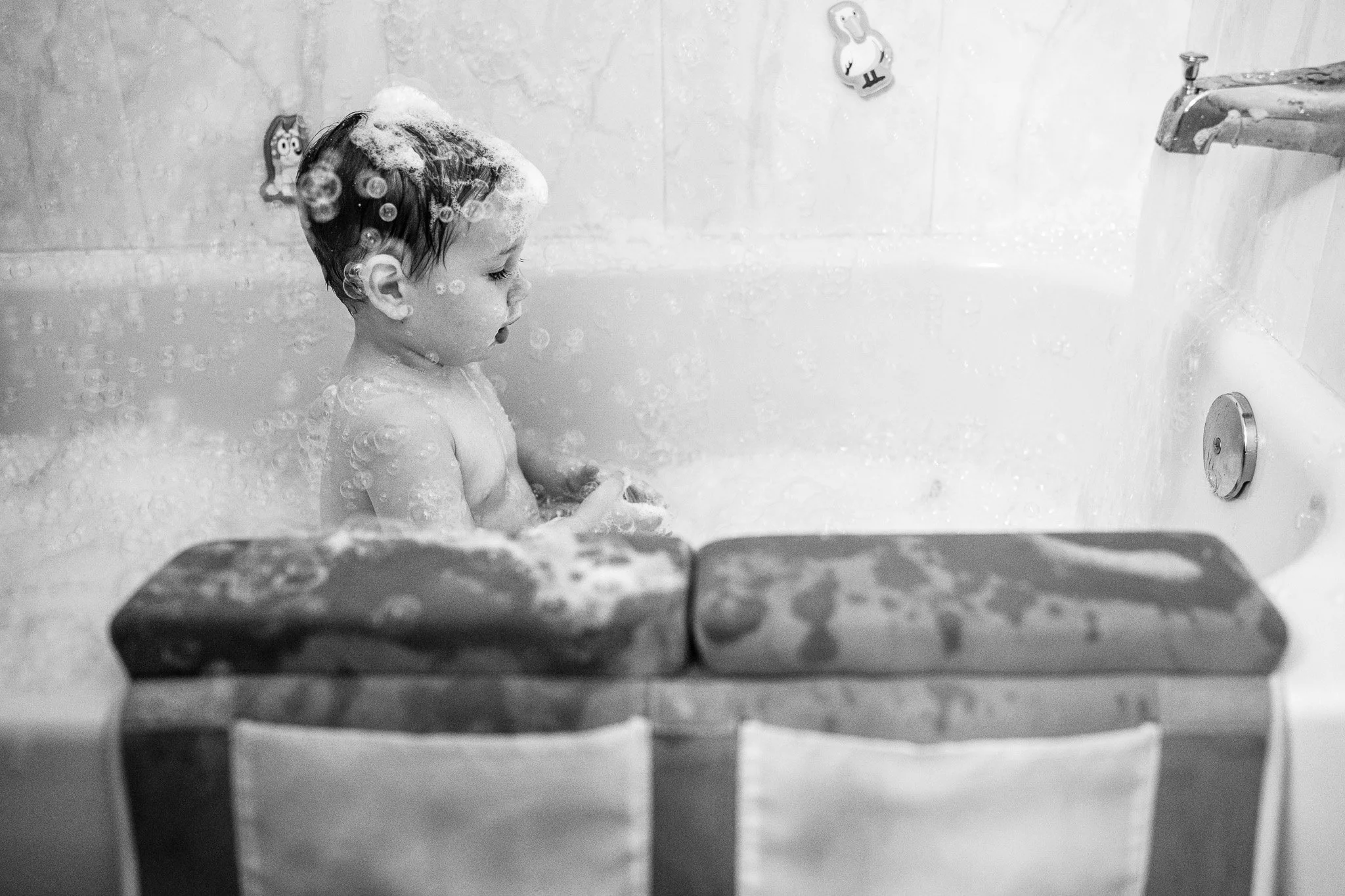 A young child taking a bath in a bathtub with soap bubbles on the head, looking down with a peaceful expression. The bathtub has a faucet on the right side, and there are children's bath toys attached to the wall.