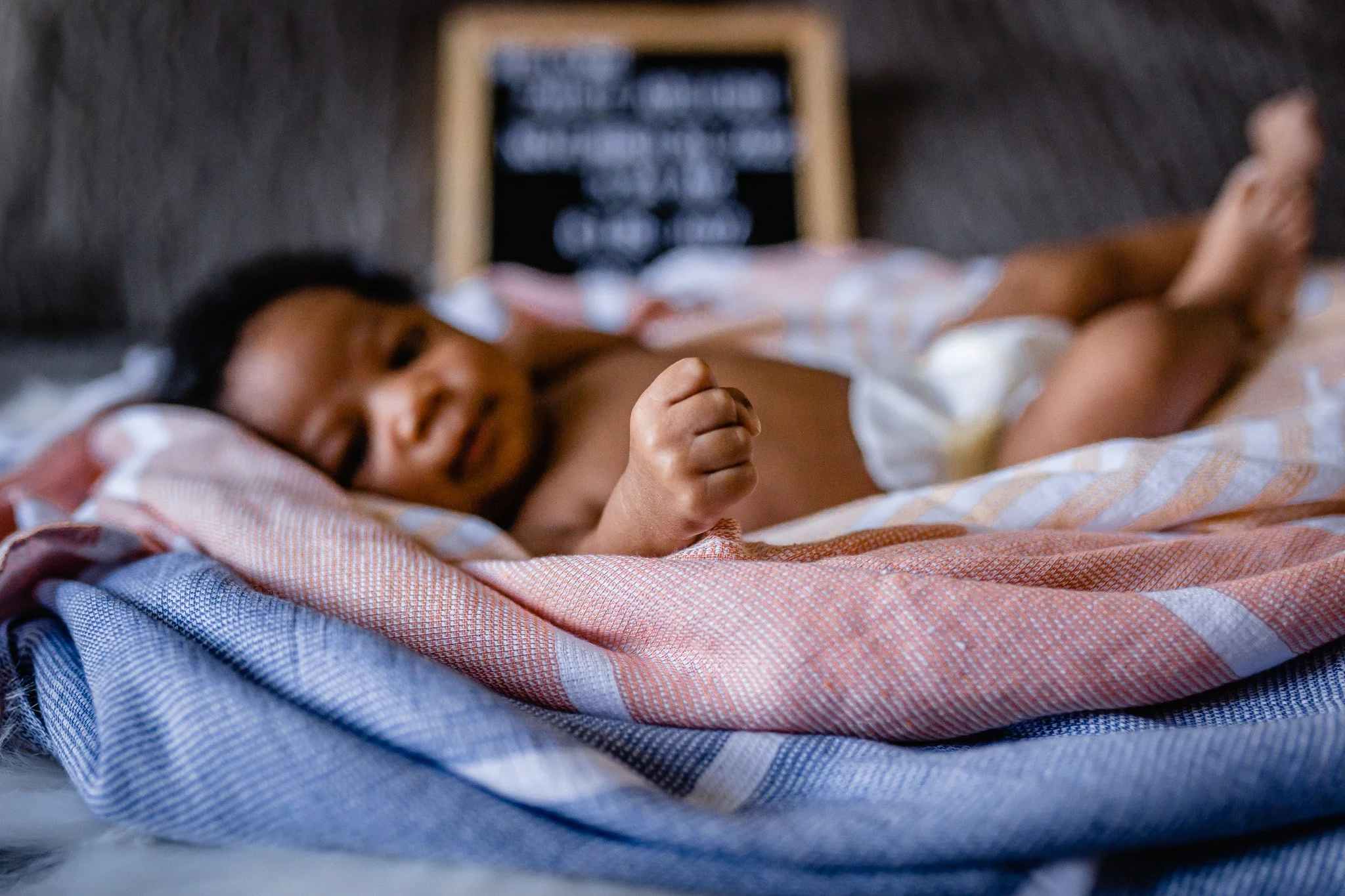 A baby lying on a bed covered with striped blankets, looking at the camera with a slight smile, with a chalkboard in the background.