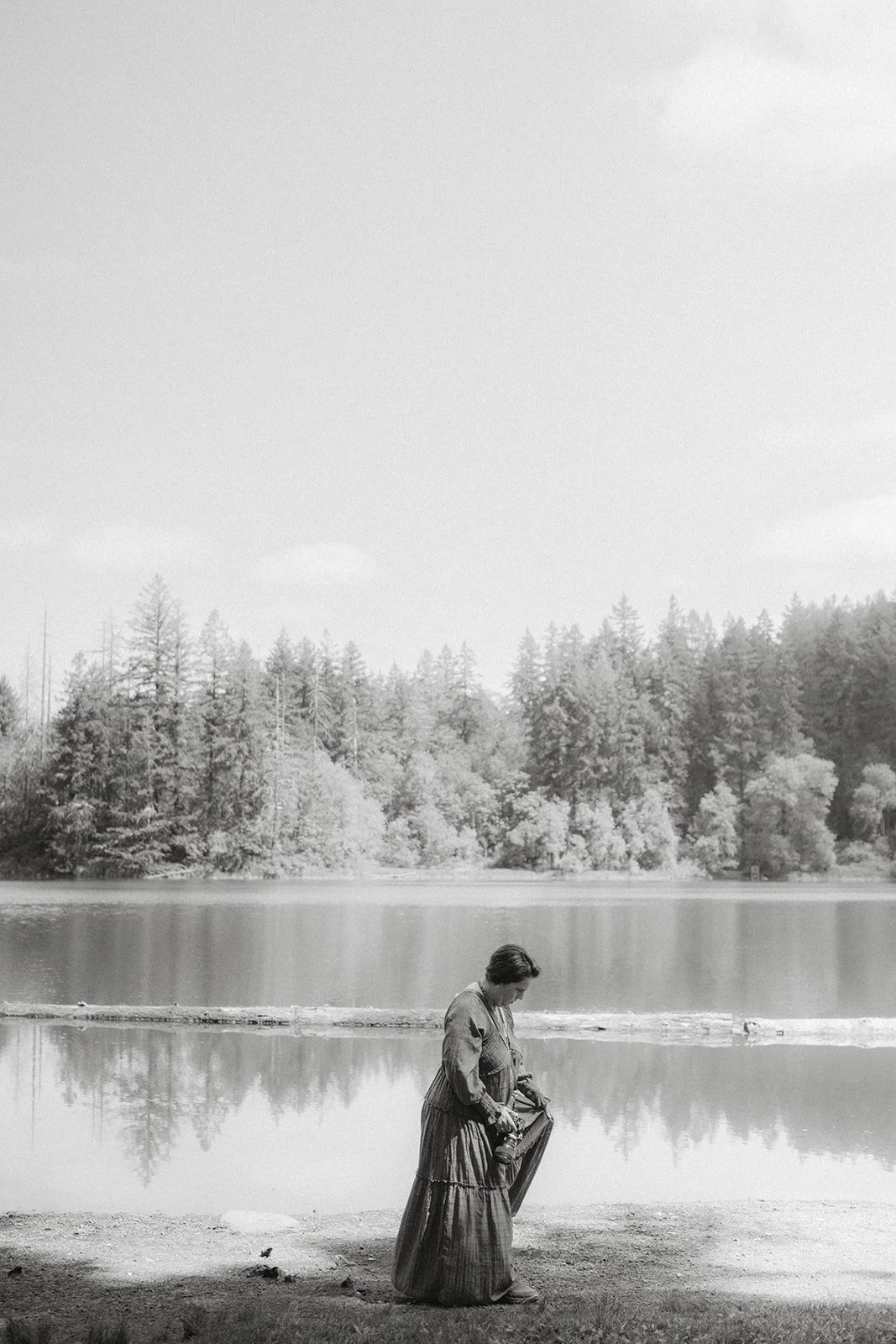 Black and white photo of a woman standing by a lake, holding her dress, with trees and a cloudy sky in the background.