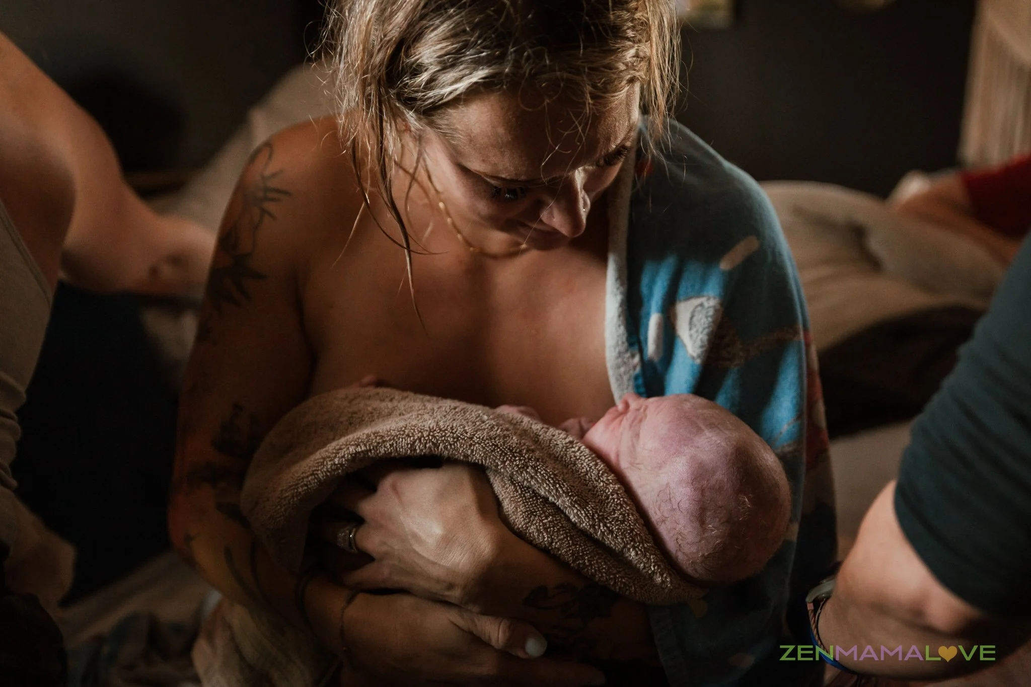 A woman with tattoos on her arm and blonde hair bends over a newborn baby wrapped in a towel, looking at the baby with affection. The scene appears warm and intimate, likely a birth or post-birth moment.