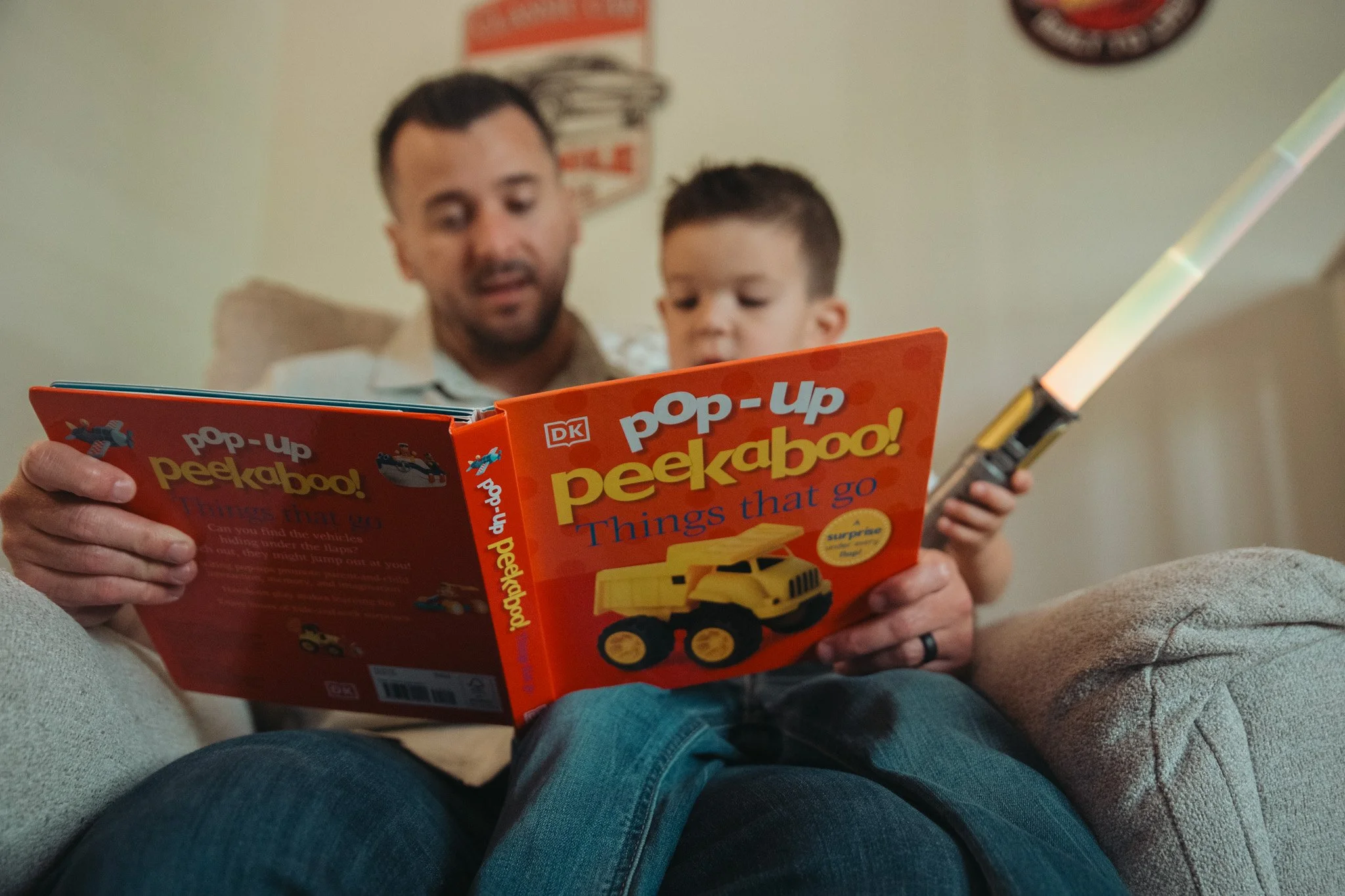 A man with a child sitting on a couch, reading a bright orange kids' book titled 'Pop-Up Peekaboo!'. The man has short dark hair and a beard, and the child has short brown hair. The background has a green wall.