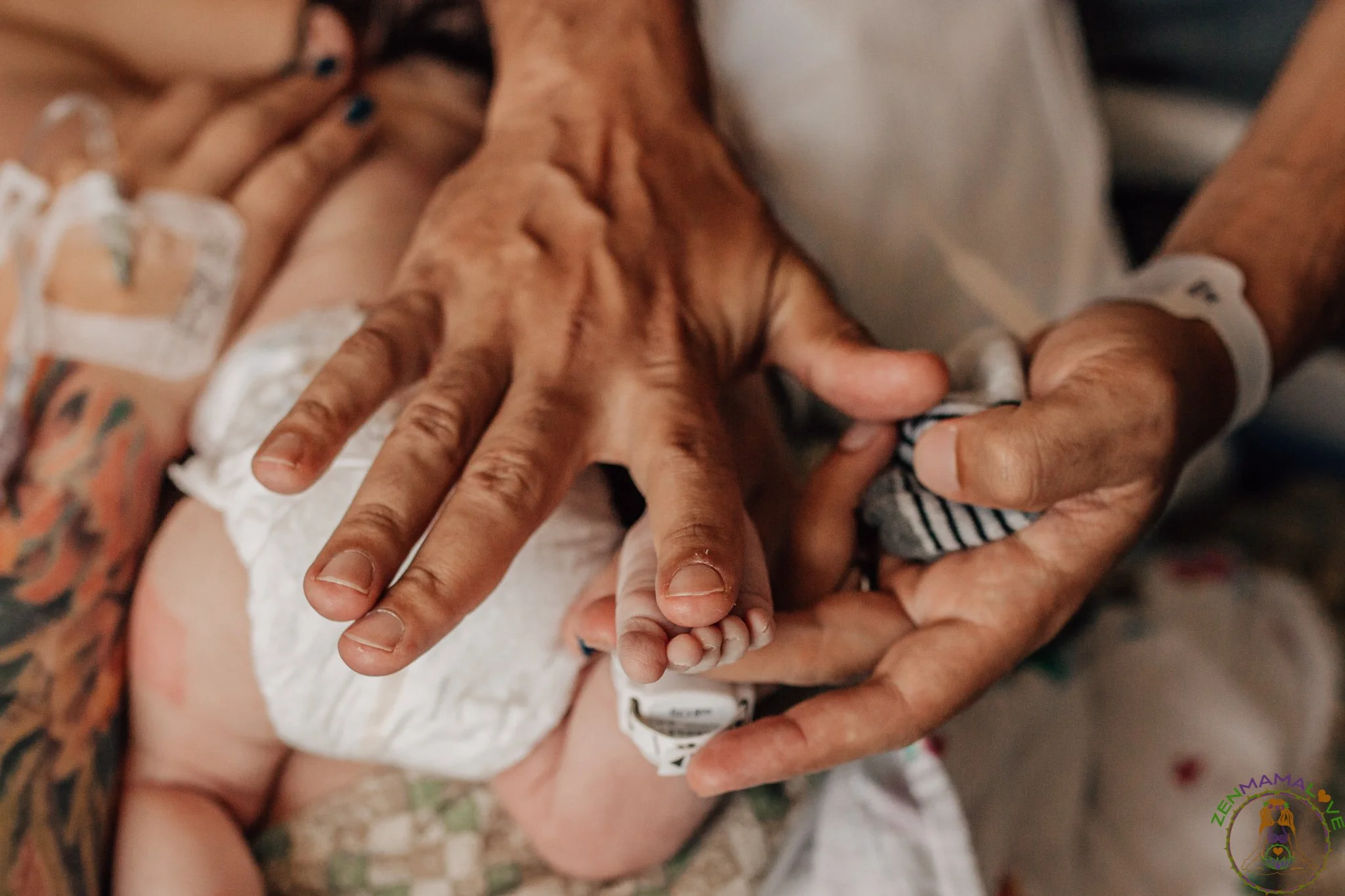 A close-up of multiple hands, including a newborn baby's tiny hand reaching out, held together in a display of unity and support. The background shows a person with tattoos and a white hospital wristband, indicating a hospital or medical setting.