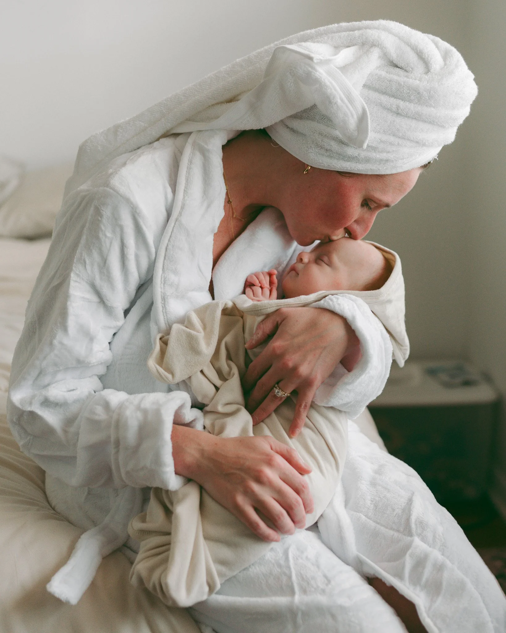 A woman wearing a white towel turban and white loungewear, cradling and kissing a sleeping baby with a beige hoodie, against a plain background.