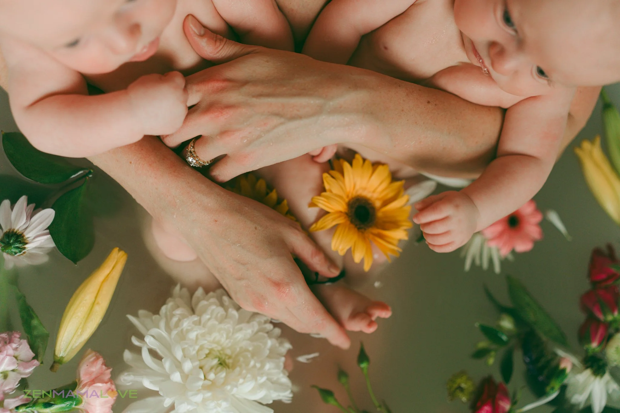 Close-up of a woman holding a baby, surrounded by colorful flowers including yellow sunflowers, white daisies, and pink blossoms.