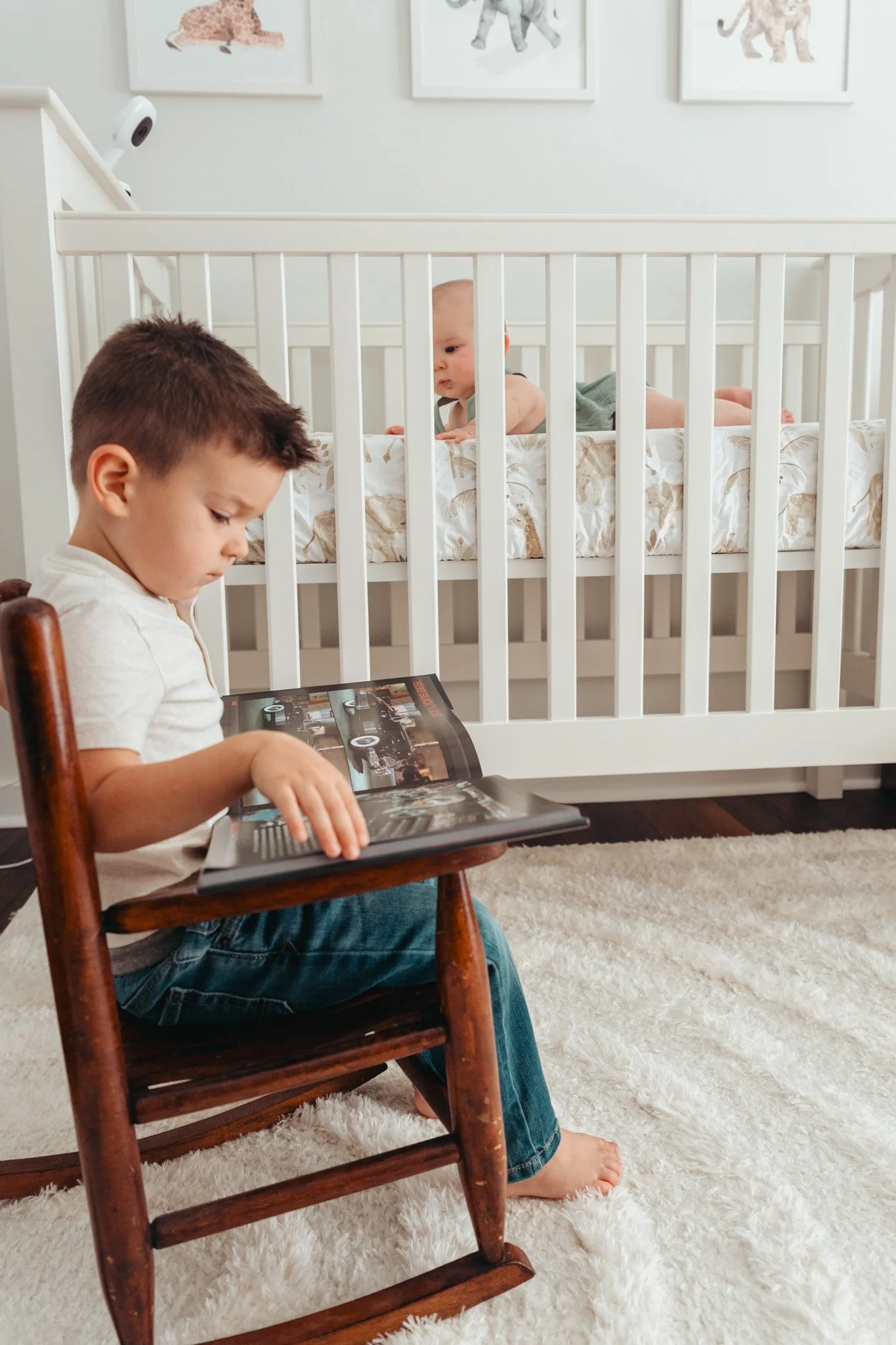 A young boy sitting on a wooden chair, reading a book, with a baby in a crib behind him looking over the bars.