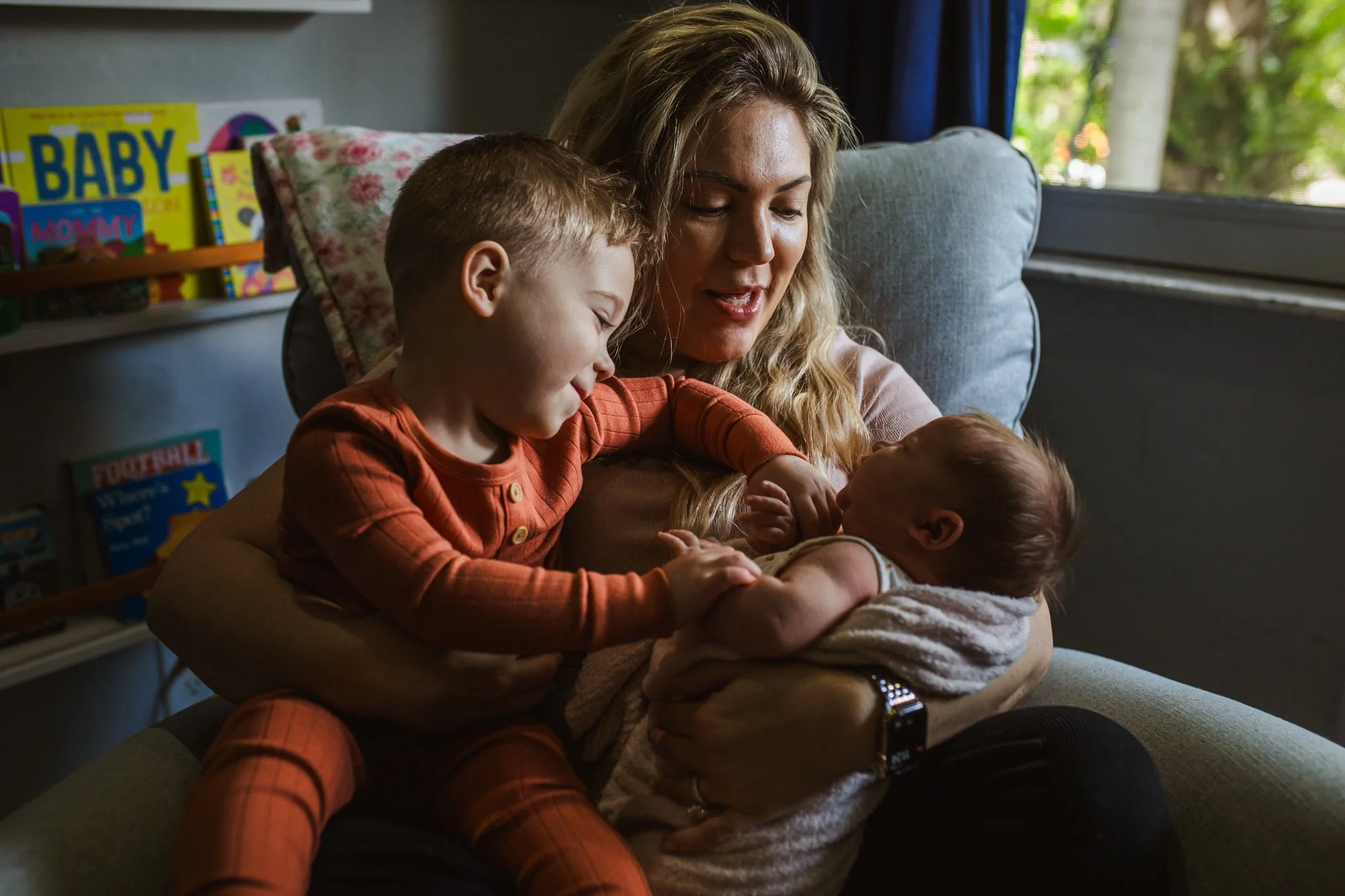 A woman with long blonde hair holding two children, one older child with short light brown hair wearing orange pajamas, and a baby wrapped in a blanket. They are sitting on a gray sofa near a window with trees outside.