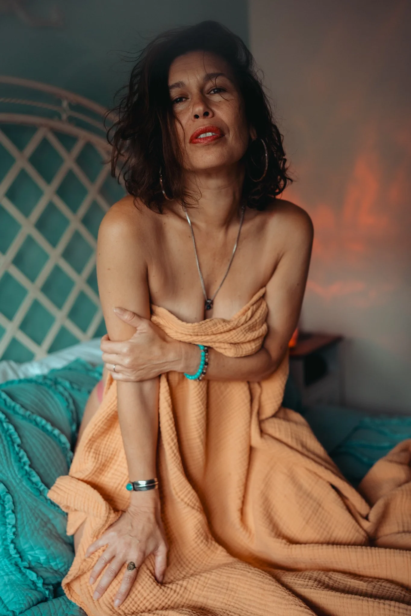 A woman with dark, shoulder-length, curly hair and wearing large hoop earrings, gold lipstick, and a teal beaded bracelet, sitting on a bed wrapped in an orange towel, with her arms crossed and looking at the camera in a room with a teal bedspread an