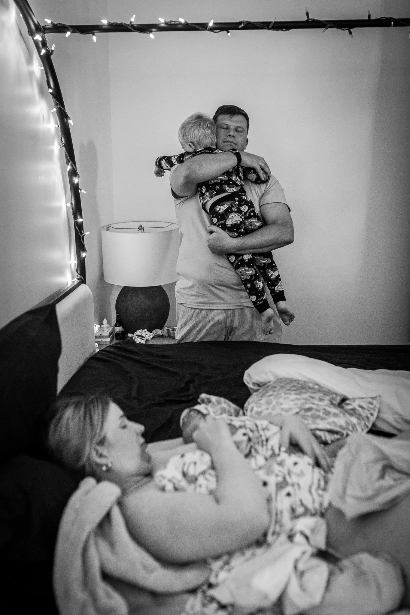 A black and white photograph of a bedroom. In the foreground, a young girl with light hair is lying on the bed, resting her head on a pillow, and covered with a blanket. In the background, a man is holding a young child and hugging them with eyes clo