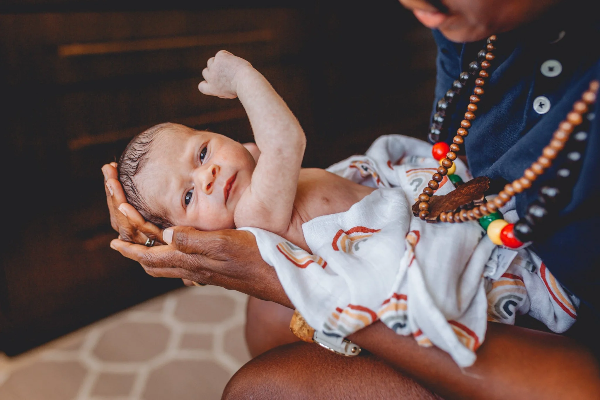 A young infant with light skin and dark hair lying in someone's arms, looking up with a calm expression. The person holds the infant's head and chest, and the infant is dressed in a colorful patterned shirt. The adult is wearing a blue shirt and a be