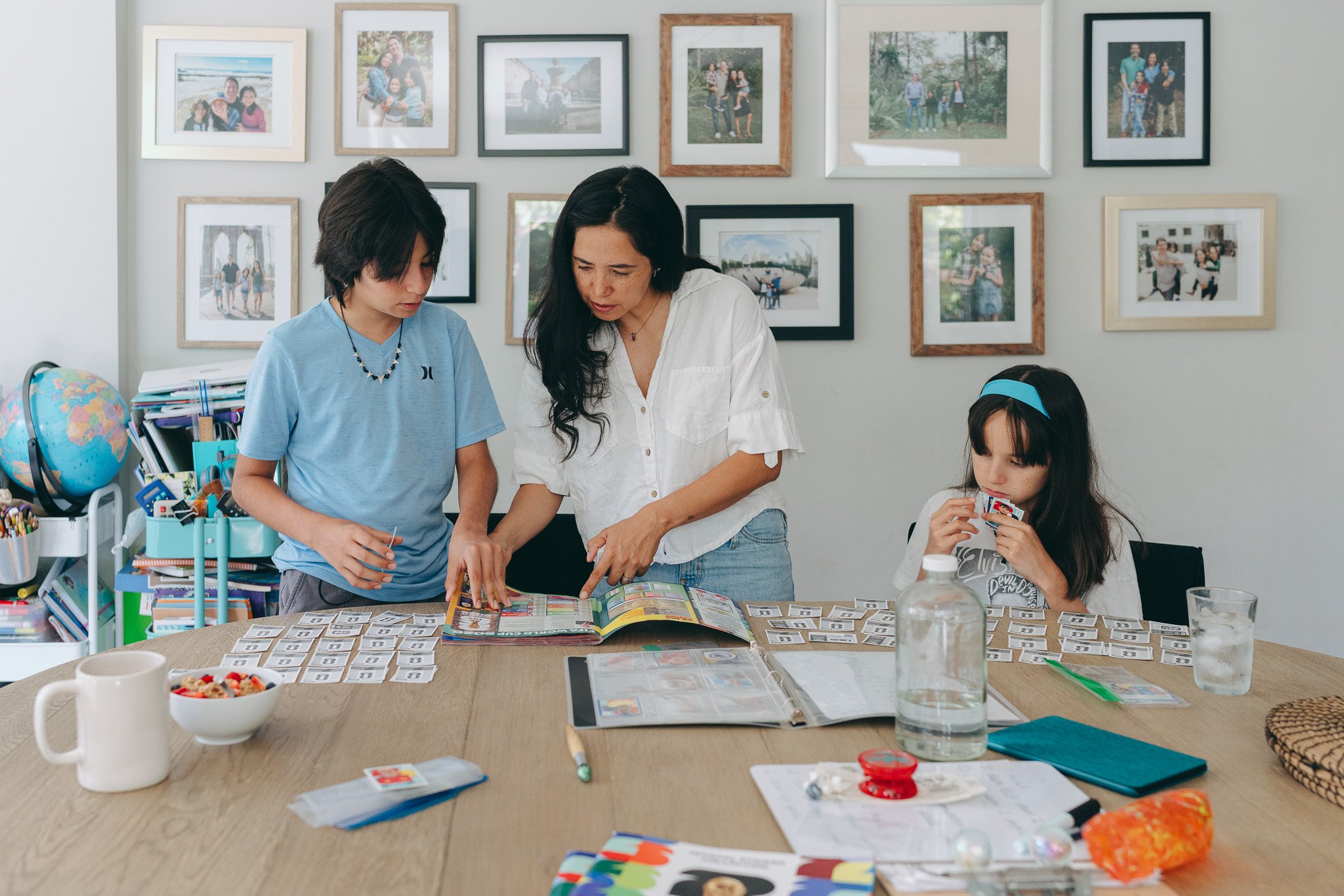 A woman and two girls sitting at a table with various educational materials, playing a matching card game. The background features a wall decorated with numerous framed family photos. The table also has a globe, a bowl of snacks, water bottles, and o