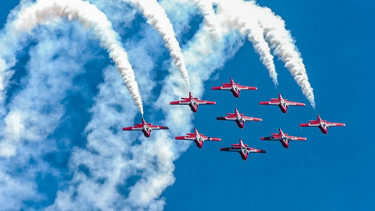 Aerial performance with nine red jets in formation, releasing white smoke trails against a blue sky.