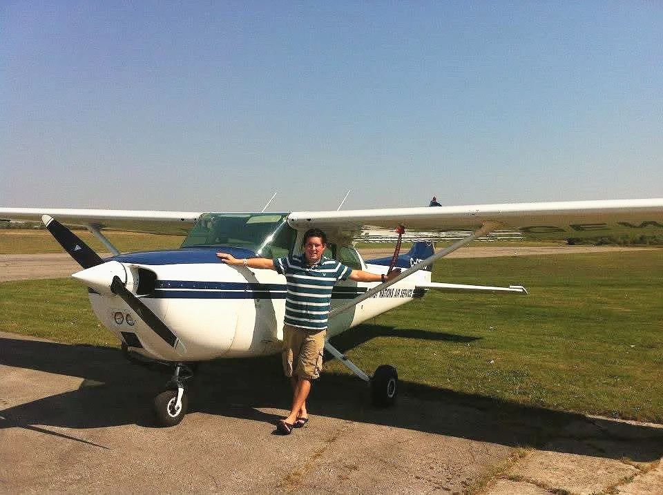 A person standing in front of a small private airplane, posing with arms outstretched outdoors on a clear day.