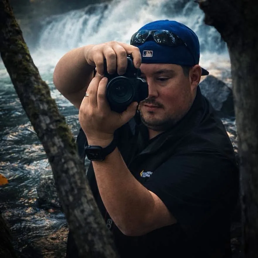 Man in a black shirt and blue cap taking a photograph with a camera near a waterfall, surrounded by tree branches.