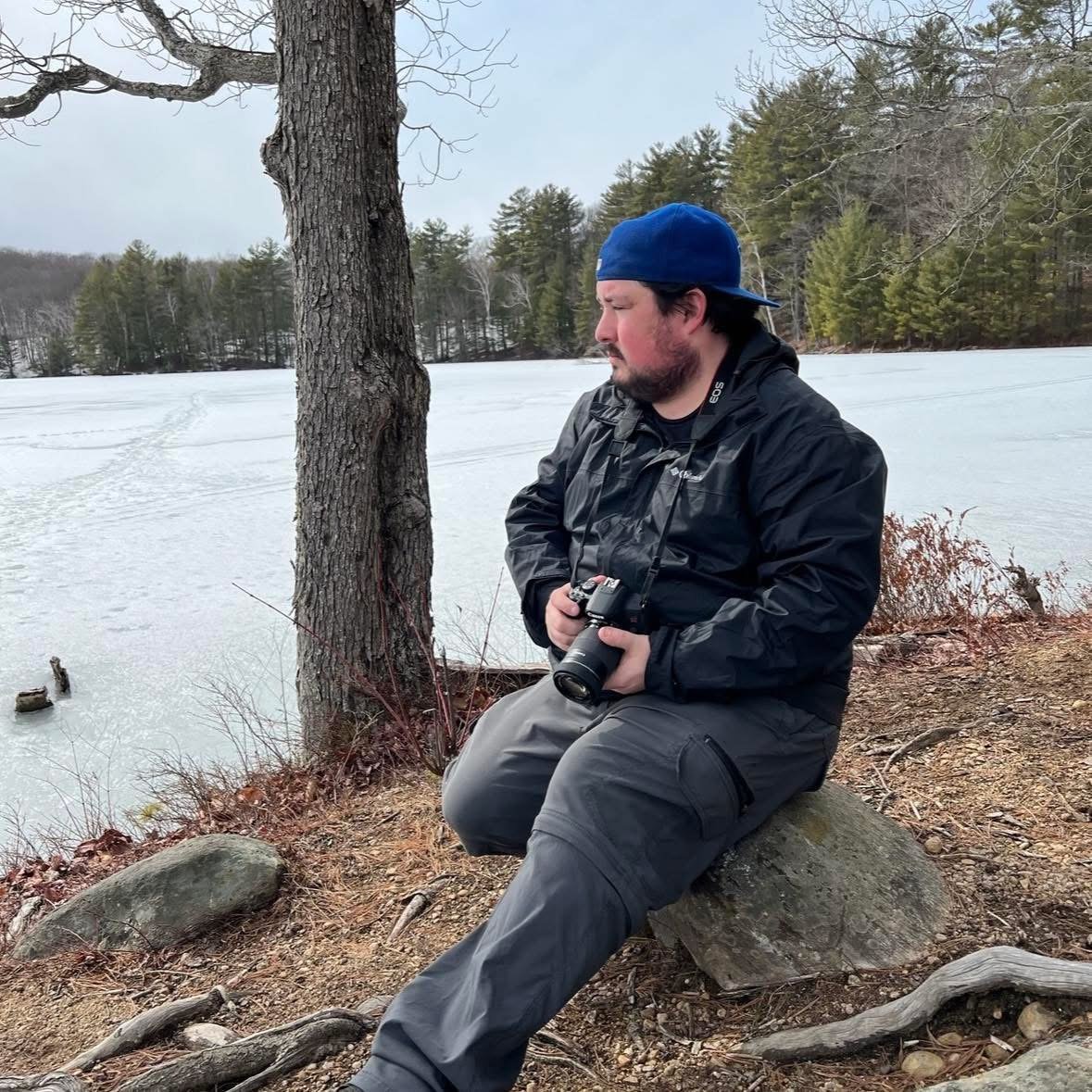 A man sitting on a rock by a frozen lake, holding a camera with a winter forest in the background.