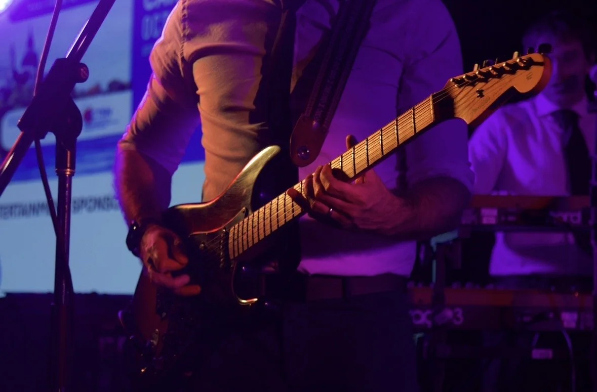 Person playing an electric guitar on stage with purple and blue lighting, with a keyboard player in the background.