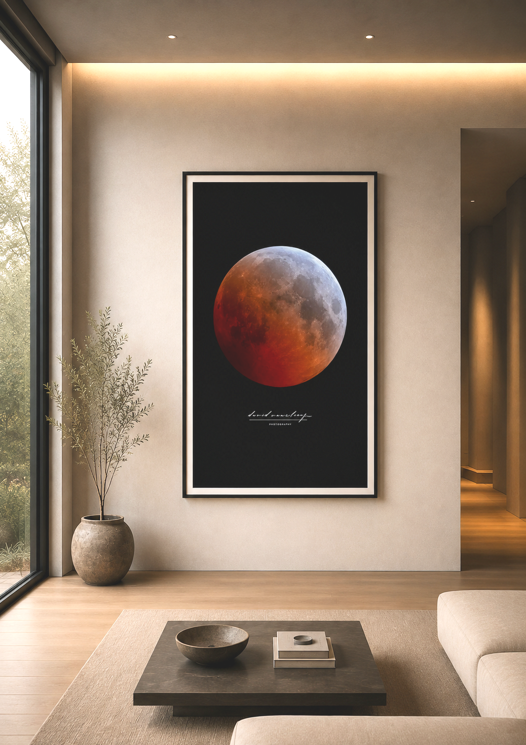 A modern living room features a large framed poster of a blood moon on a black background, with alcove lighting above. There is a potted plant with thin branches in a round concrete pot next to a floor-to-ceiling window on the left, and a sofa and coffee table with decorative items in the foreground.