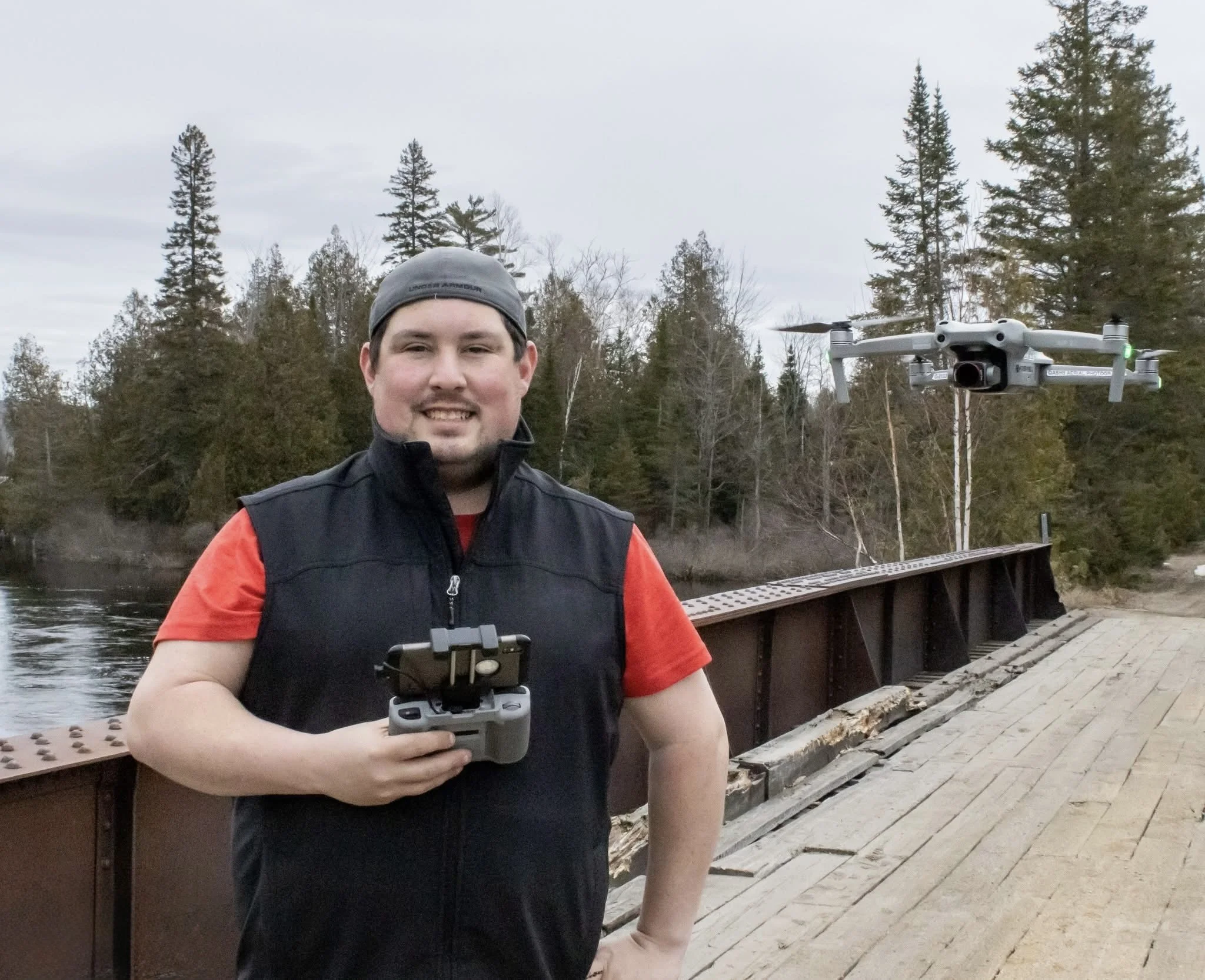A man wearing a gray cap, black vest, and red shirt standing on a wooden bridge outdoors with a drone flying nearby and trees in the background.