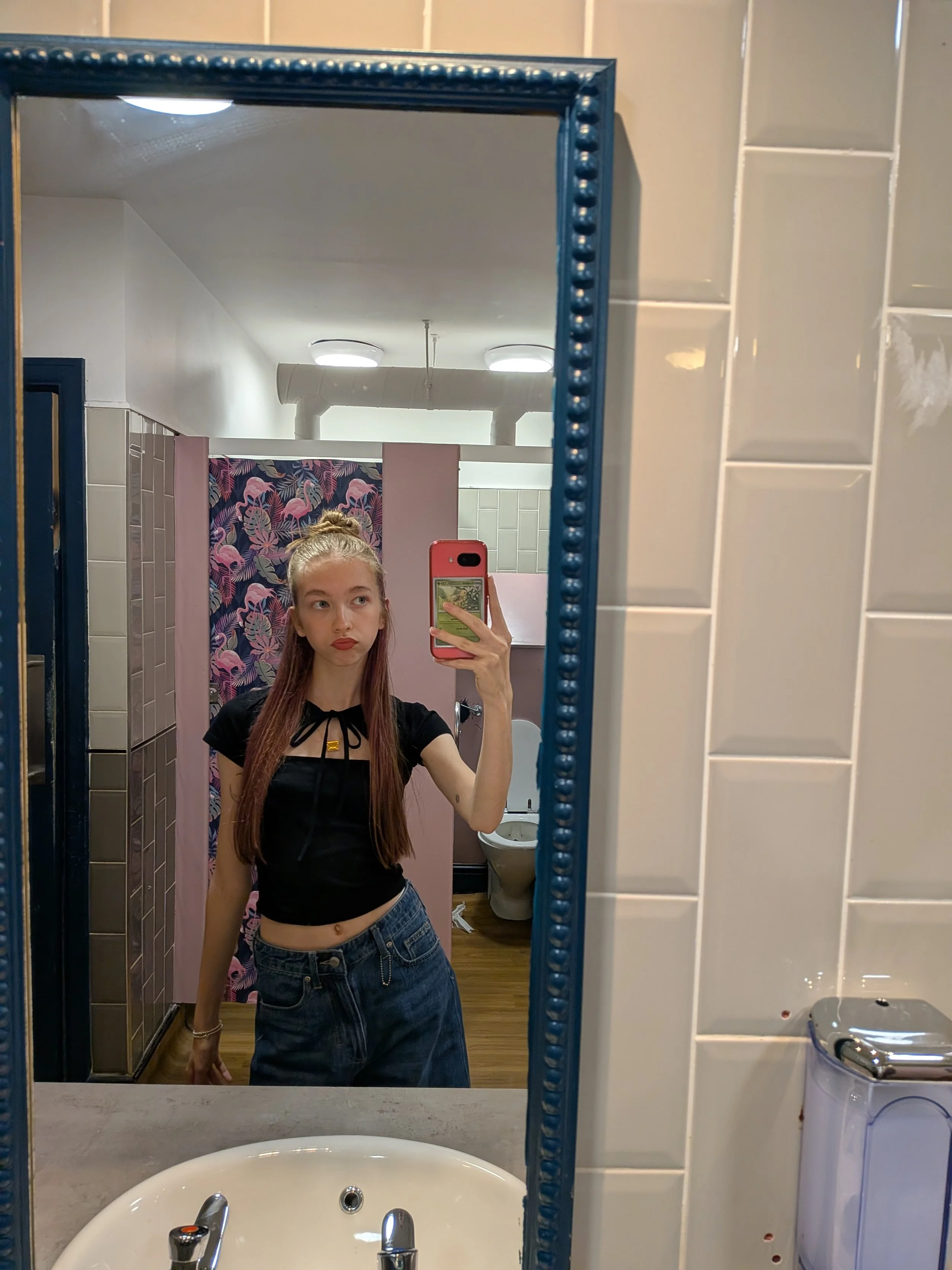 A young woman with long hair tied in a bun taking a mirror selfie in a bathroom. She wears a black top with a lace-up front and high-waisted shorts. The bathroom has a decorative mirror with blue beaded trim, a pink shower curtain with flamingo and leaf patterns, and a white tiled wall. A toilet and washing machine are visible in the background.