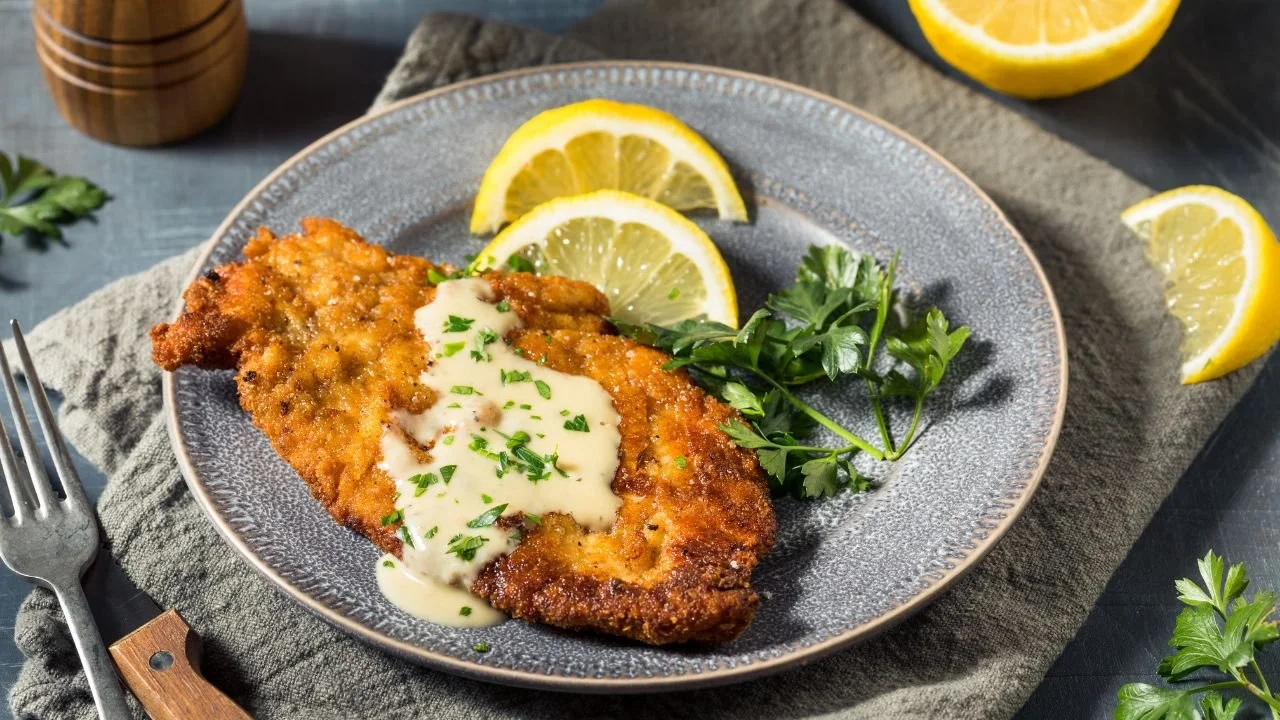 A plate of breaded, fried fish fillet topped with creamy white sauce, garnished with chopped parsley, served with lemon wedges and a sprig of parsley on a gray plate.