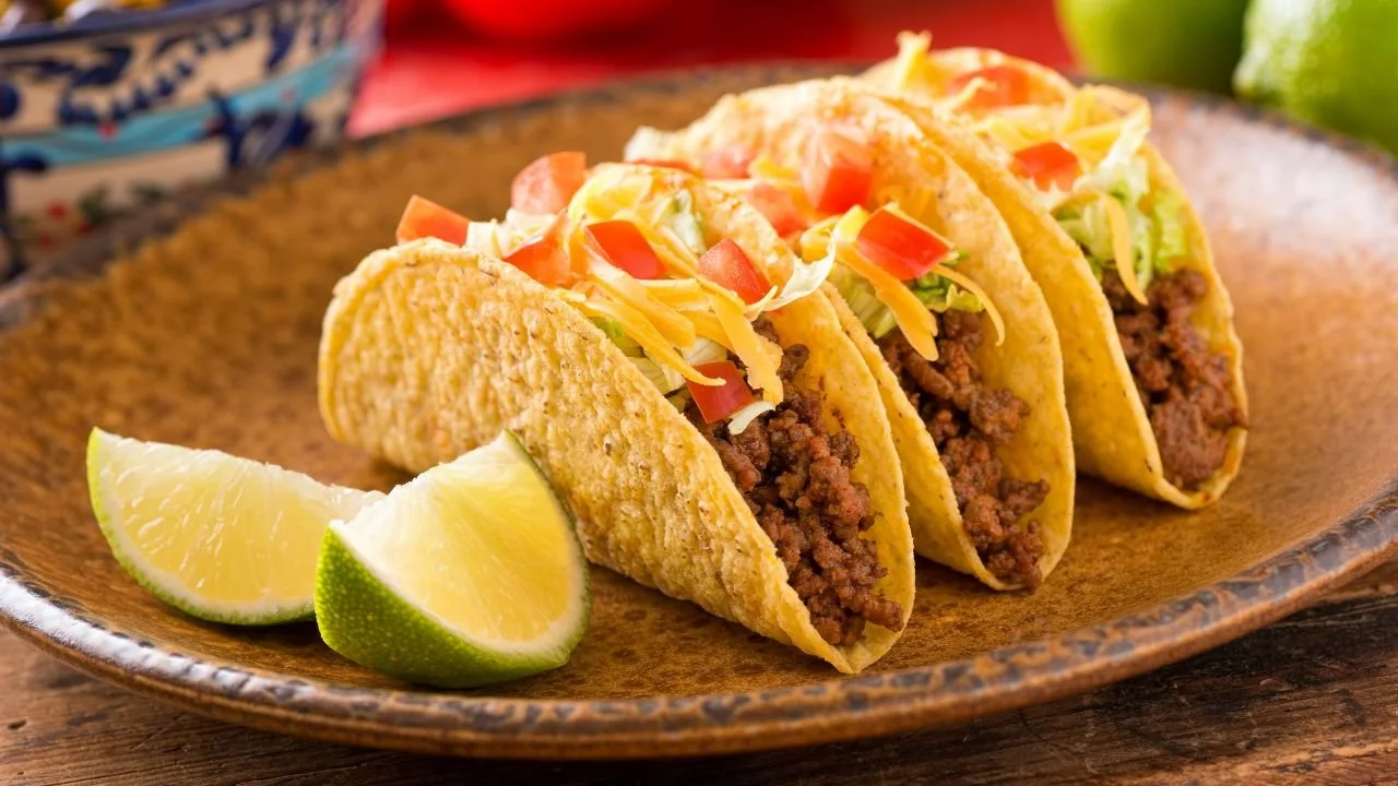 Three soft tacos filled with seasoned ground beef, shredded lettuce, diced tomatoes, and shredded cheese, served on a wooden plate with lime wedges.