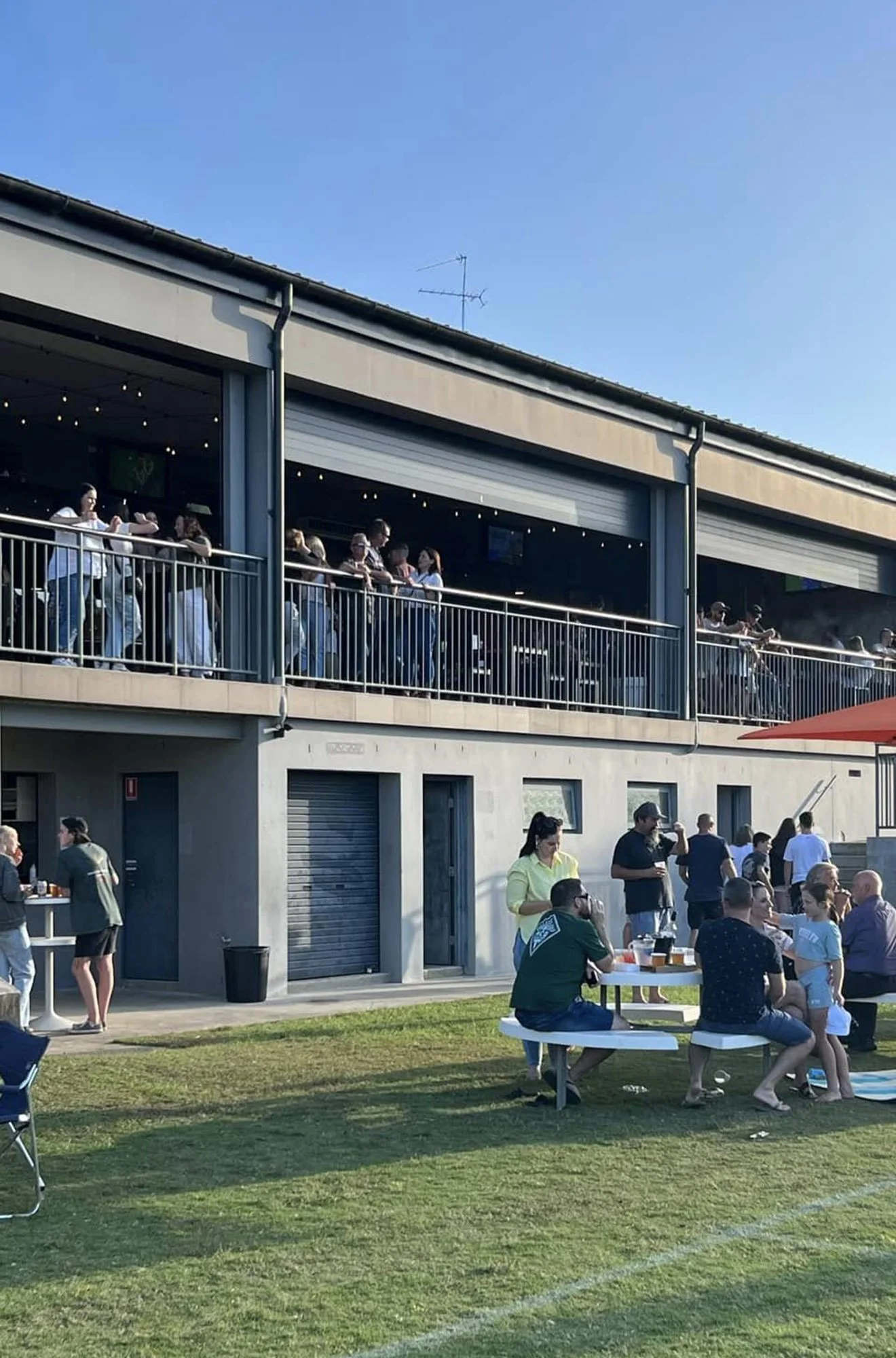 People gathering outside a two-story building with an open balcony, some standing on the balcony and others on the lawn, enjoying food and drinks under a clear blue sky.