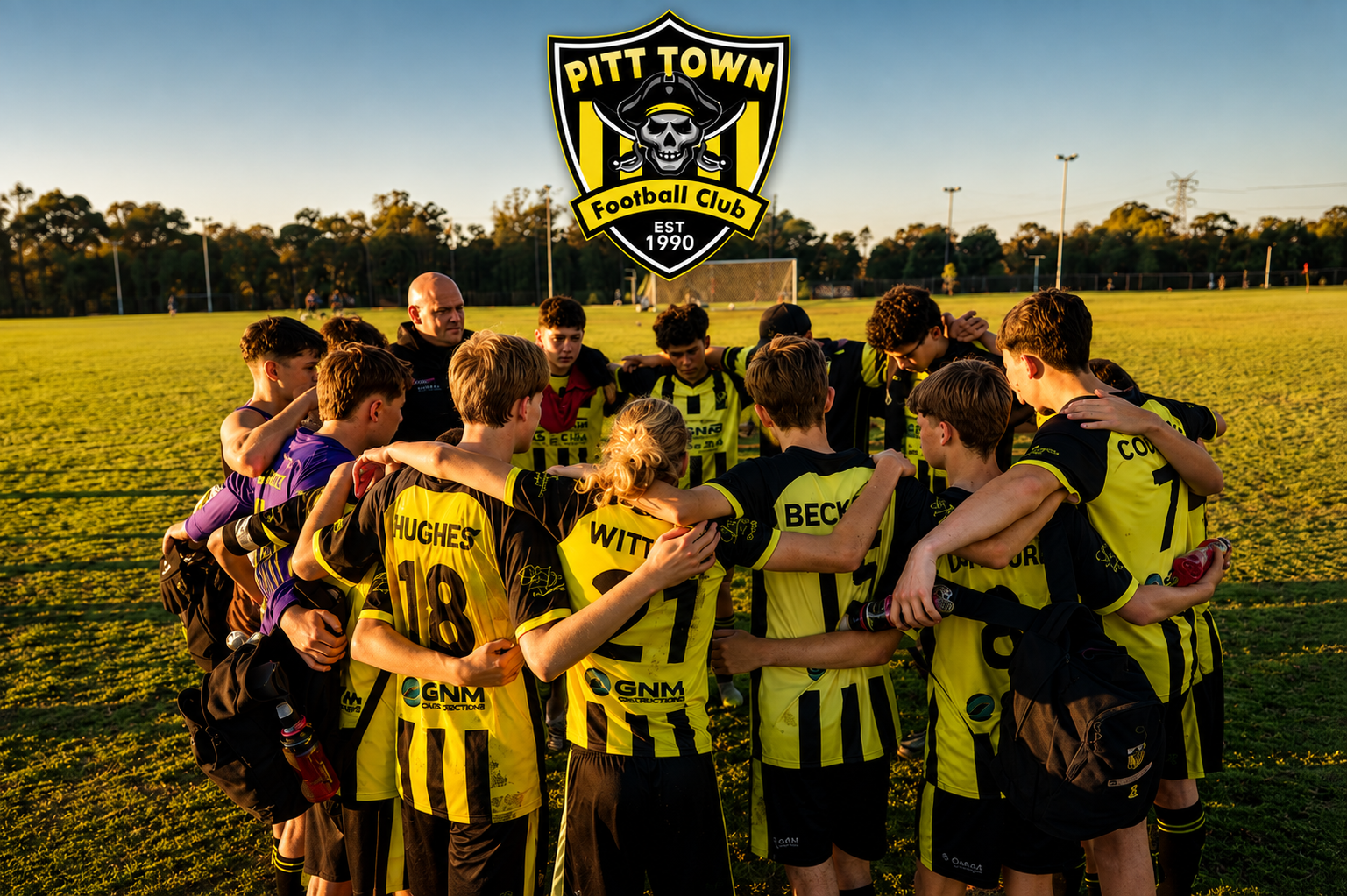 Soccer team huddle on a field during sunset with a Pitt Town Football Club crest in the sky.
