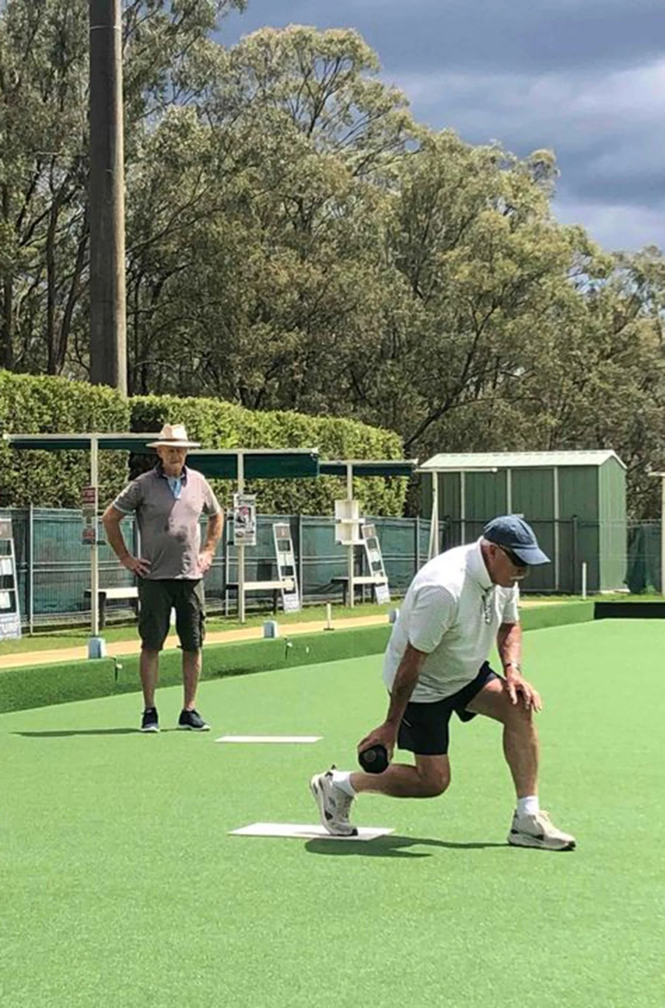 Two men playing lawn bowls on a green outdoor lawn with a hedge, trees, and a shed in the background. One man is kneeling on the ground holding a black ball, while the other man is standing nearby observing.