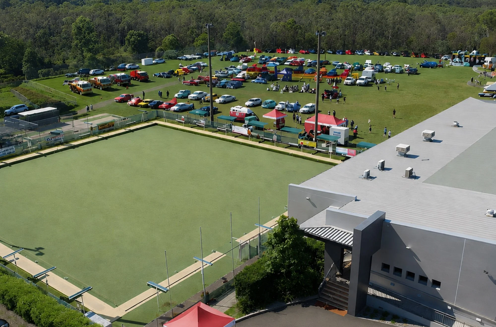 An aerial view of a sports complex with a field and a parking lot filled with cars. The parking lot is behind the fenced field, and there are colorful play structures on the grassy area. The surrounding area includes trees and hills.