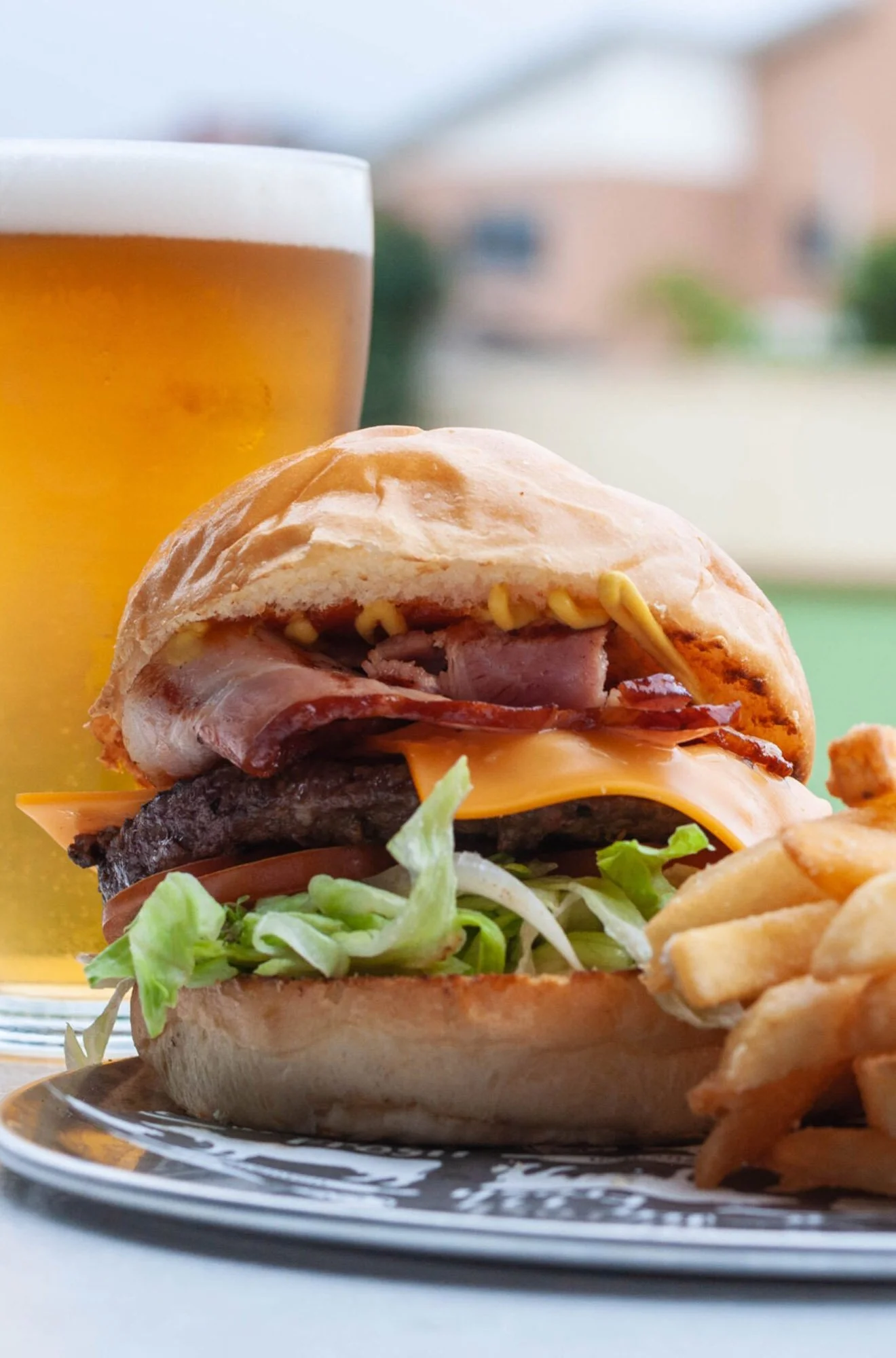 Close-up of a cheeseburger with lettuce, tomato, bacon, and cheddar cheese, served with French fries and a glass of beer in the background.