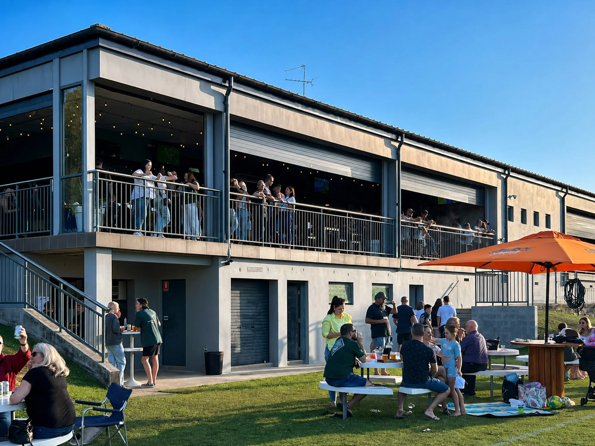 People gathered outdoors at a social event on a grassy area with tables, chairs, and an orange umbrella, beside a modern building with large windows and a balcony, under a clear blue sky.