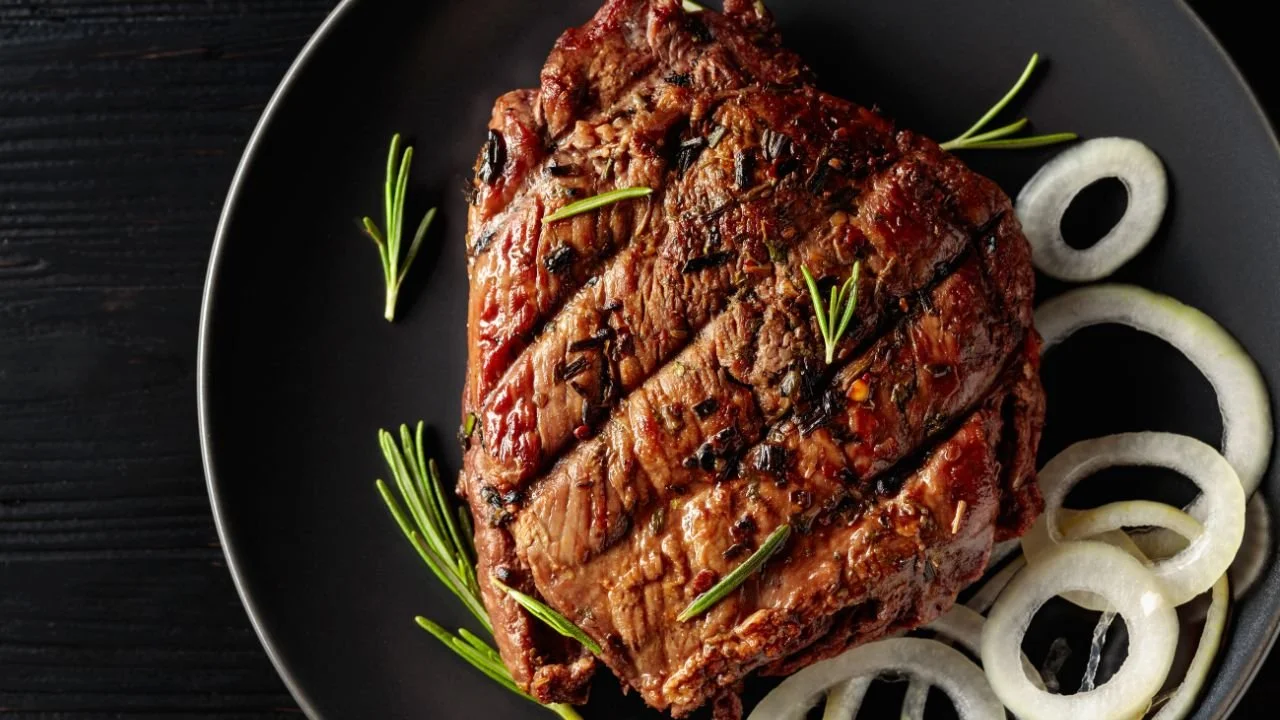 A grilled steak garnished with rosemary and sliced onions on a black plate.