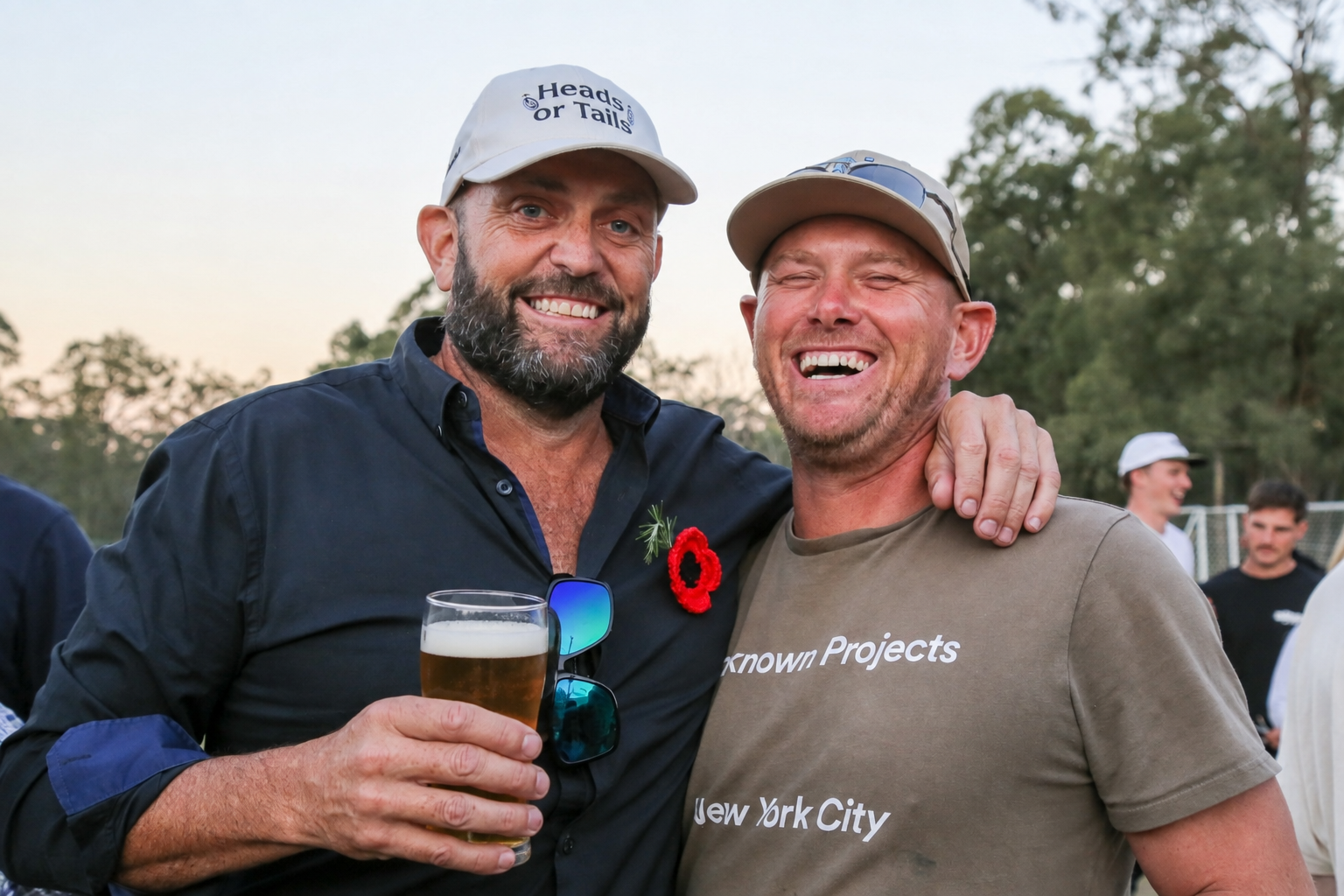 Two men smiling and hugging at an outdoor event, one holding a glass of beer and wearing a cap that says 'Heads or Tails,' the other wearing a cap and a T-shirt that says 'Known Projects New York City'.