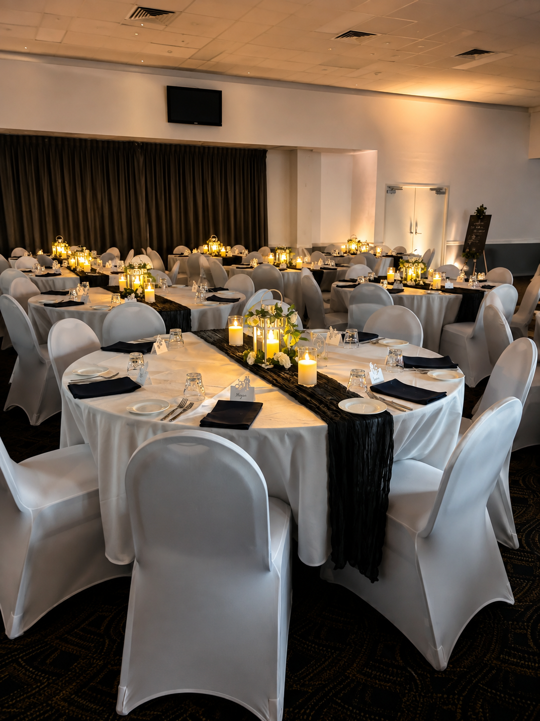 Elegant banquet hall decorated with white tablecloths, black napkins, and candle centerpieces with greenery, prepared for a formal event or wedding reception.