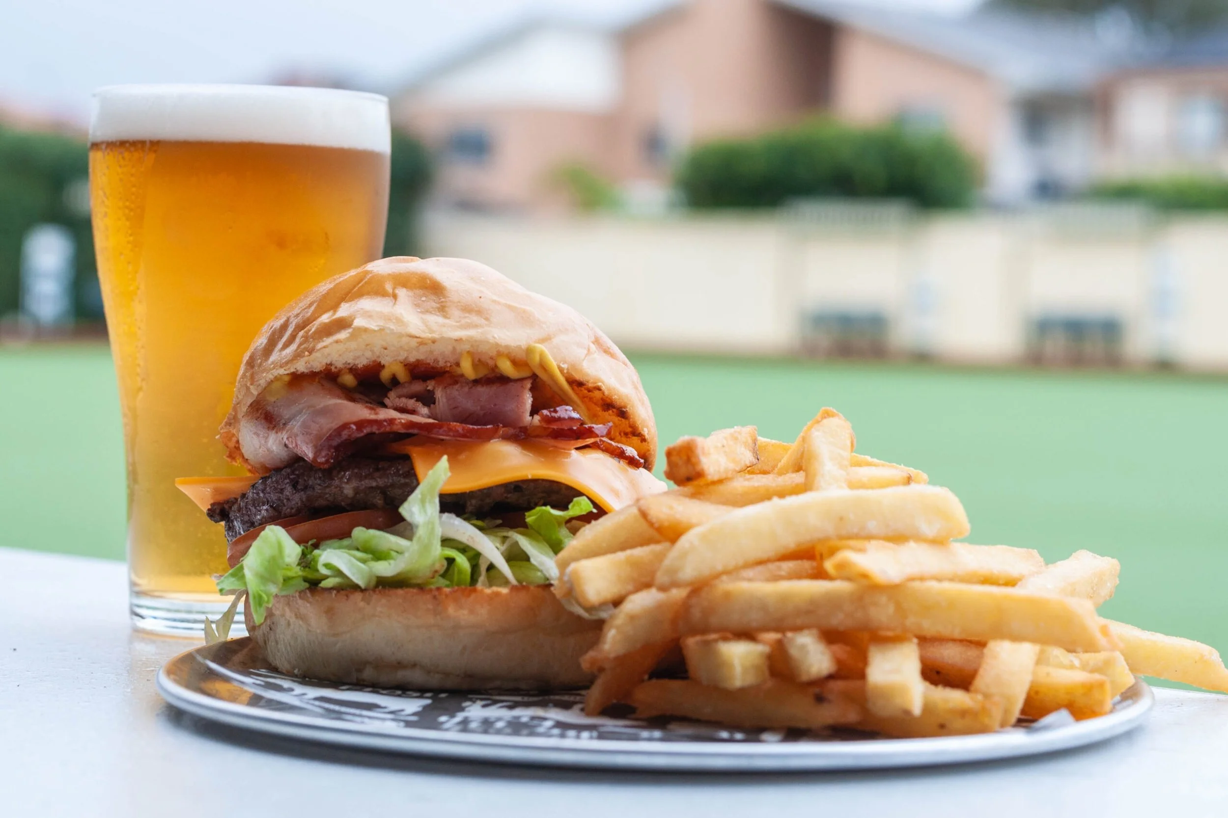 A cheeseburger with lettuce, tomato, bacon, cheddar cheese, and a burger patty in a bun, accompanied by a side of French fries, a glass of beer with foam, set outdoors with residential buildings in the background.