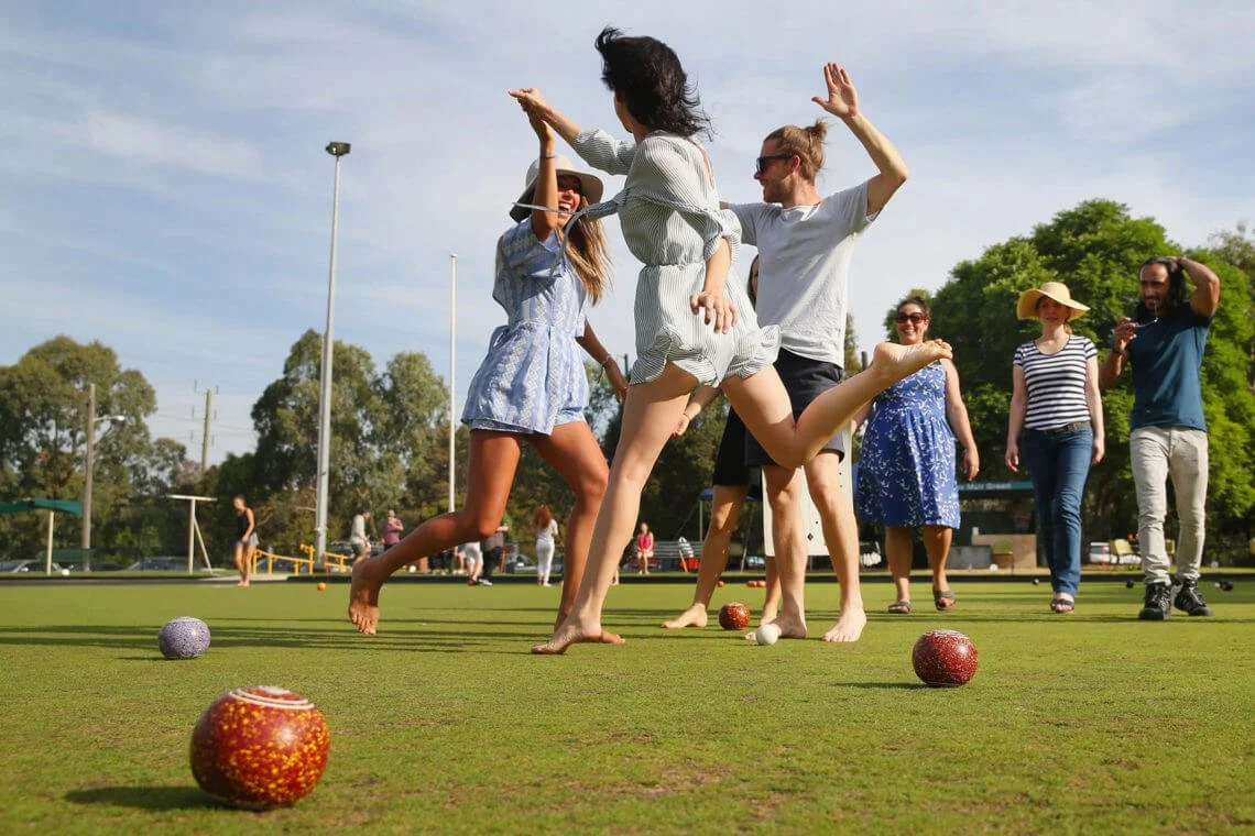 A group of people playing lawn bowls outdoors on a sunny day, with some dancers in the foreground, and others watching in the background.
