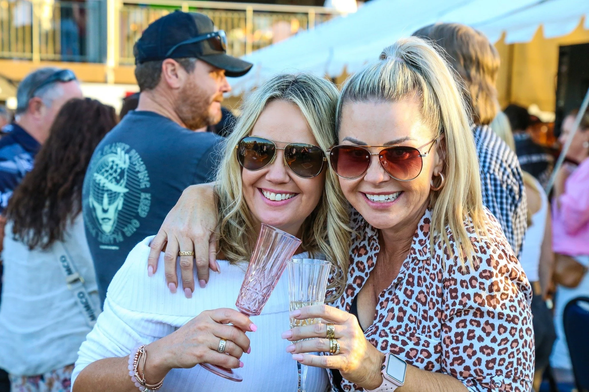 Two women with blonde hair wearing sunglasses, smiling and hugging at an outdoor gathering, holding glasses with drinks.