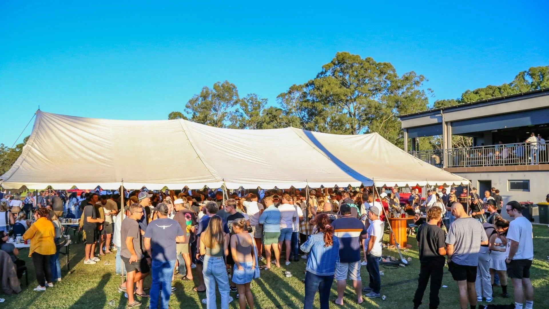 Crowd of people gathered under a large white event tent outdoors on a sunny day with clear blue sky and trees in the background.