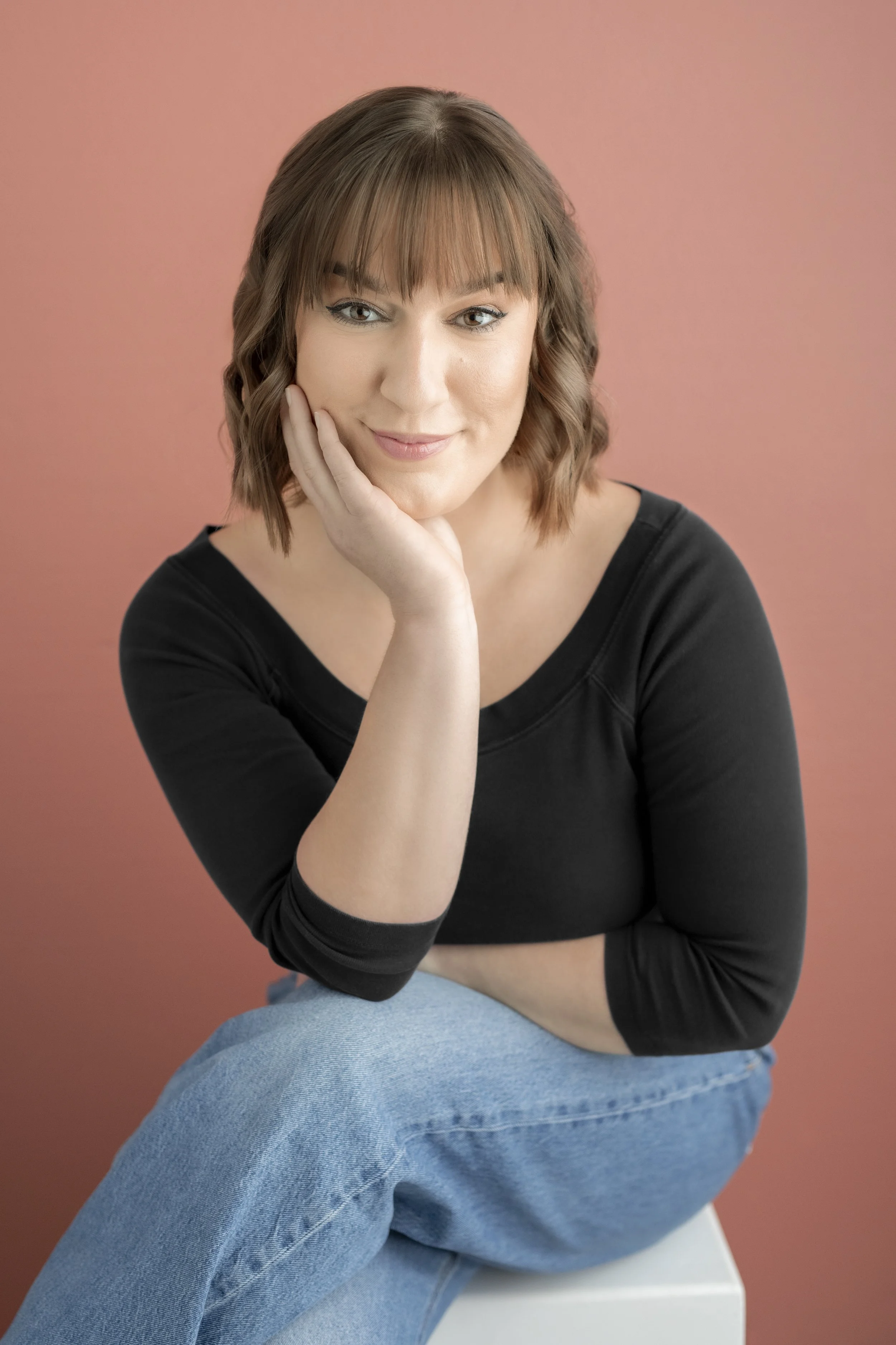A woman with brown wavy hair and bangs, smiling softly, resting her chin on her hand, wearing a black top and denim jeans, sitting against a pink background.