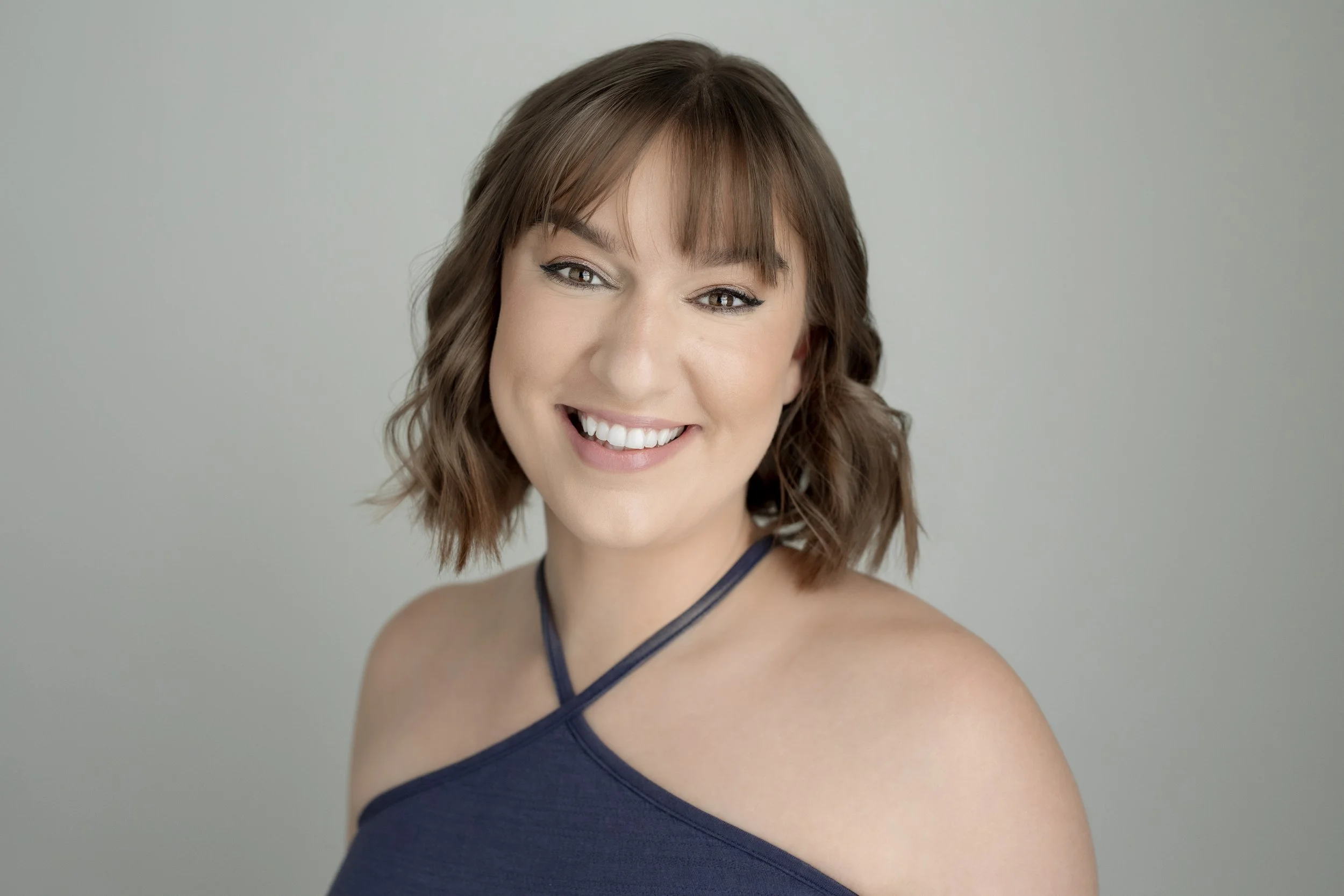 Portrait of a smiling woman with wavy brown hair, wearing a dark blue top, against a light gray background.