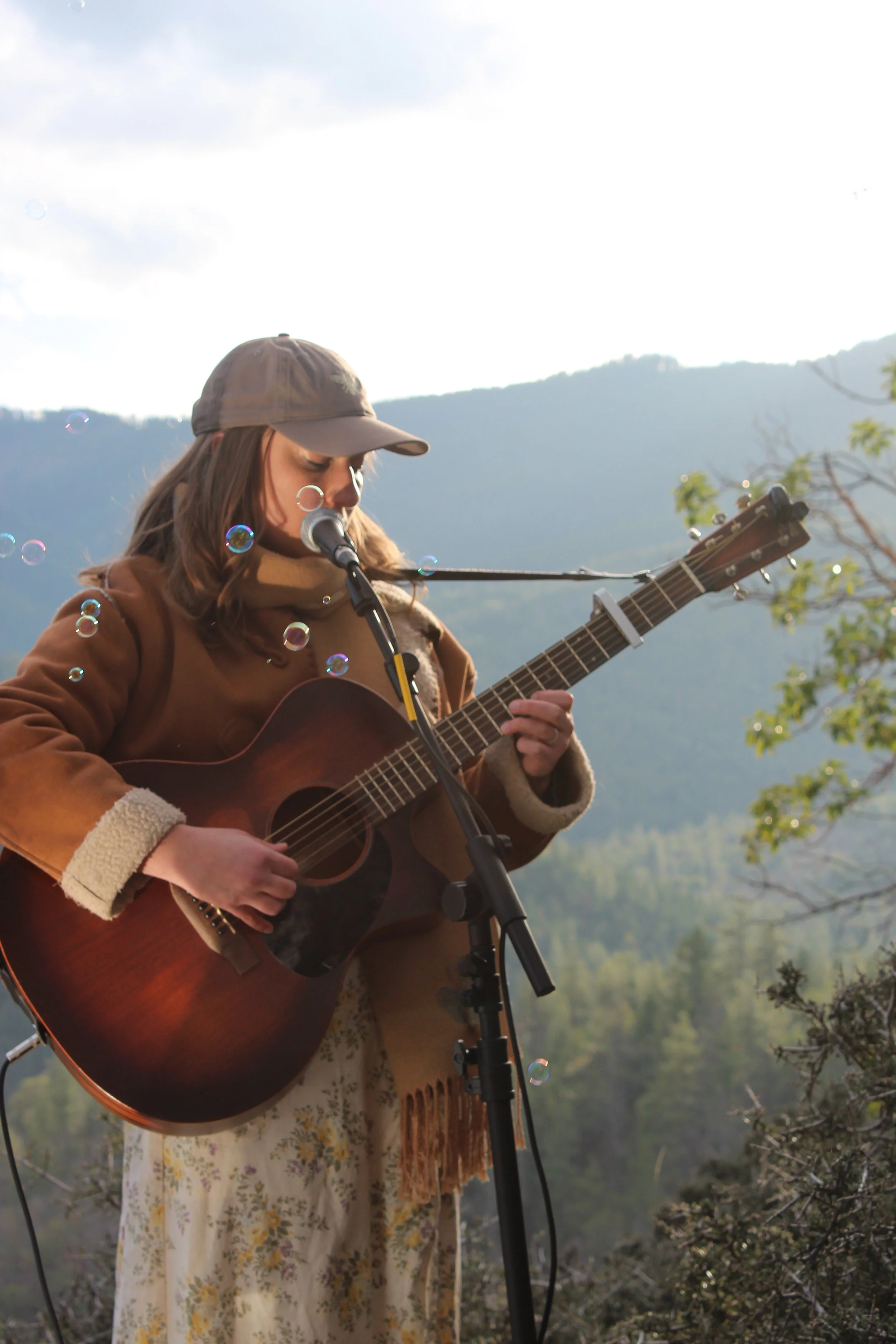 A woman wearing a tan cap, brown jacket, and floral skirt is playing an acoustic guitar and singing into a microphone outdoors with mountains in the background, with bubbles floating around.