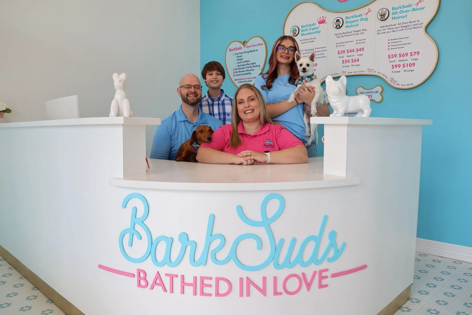The owners and their family with two dogs at the BarkSuds Sugar Land reception desk.