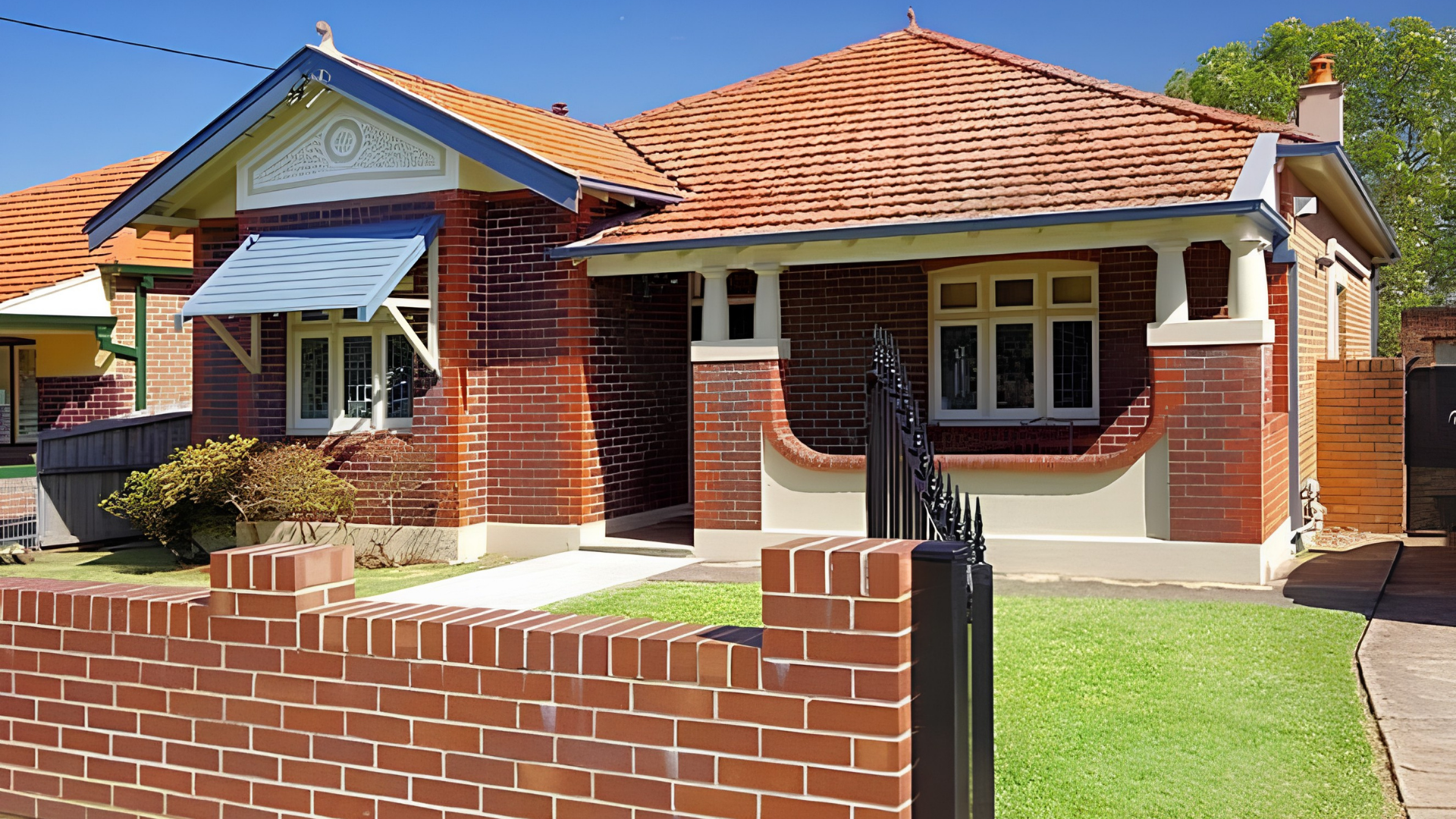 Red brick house with a gabled roof made of orange tiles, white decorative trim, and a front porch featuring white columns, a black wrought iron gate, and manicured lawn.