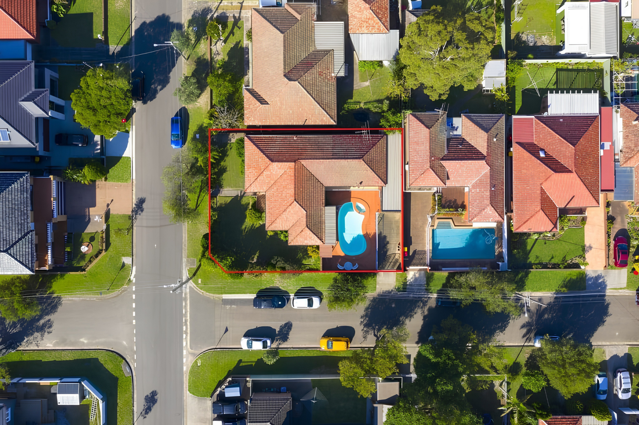 Aerial view of a residential neighborhood featuring houses with various roof styles and colors, trees, gardens, and parked cars along the streets.