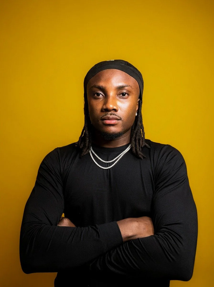 DJ Jino Spinz is shown in a Portrait  wearing a black headscarf, black long-sleeve shirt, and layered necklaces, standing against a yellow background.