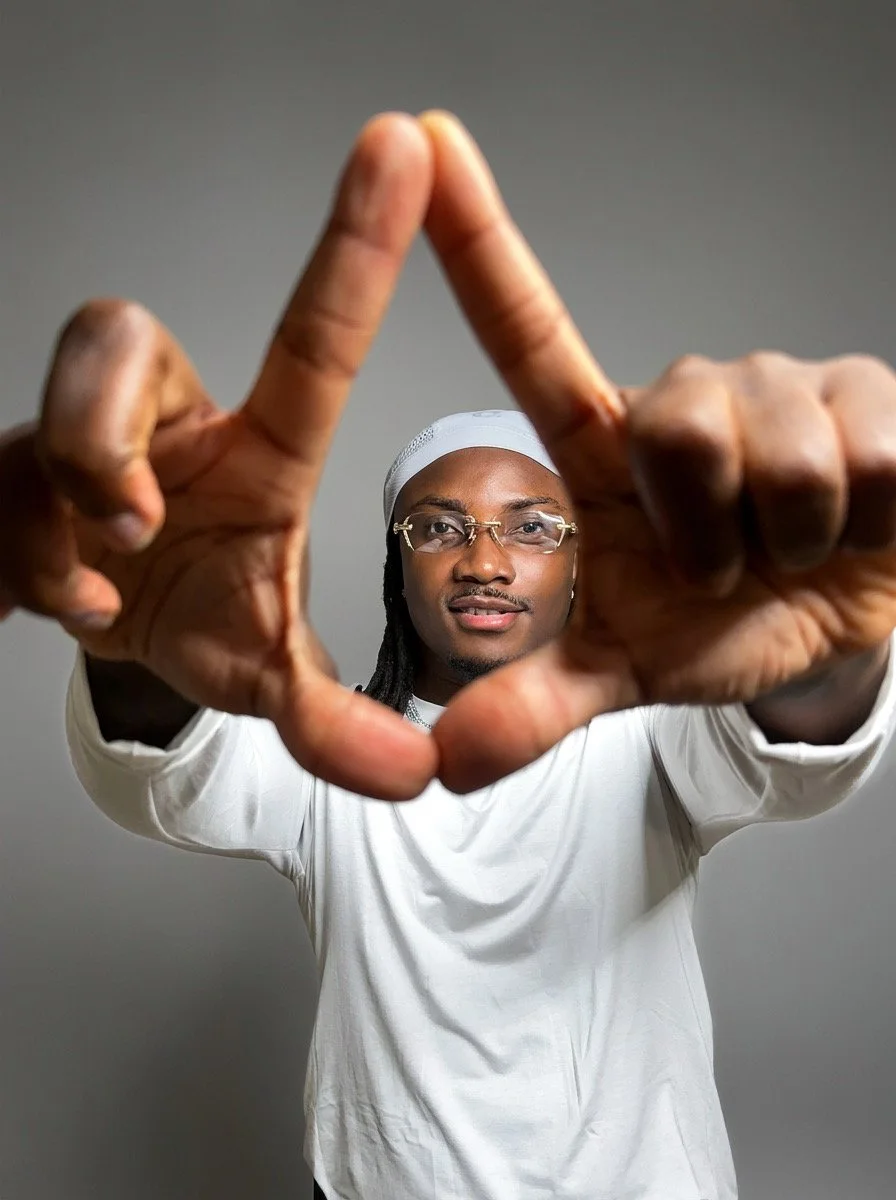 DJ Jino Spinz is wearing  glasses and white head covering forming a triangle with his hands, positioned near camera, against gray background.